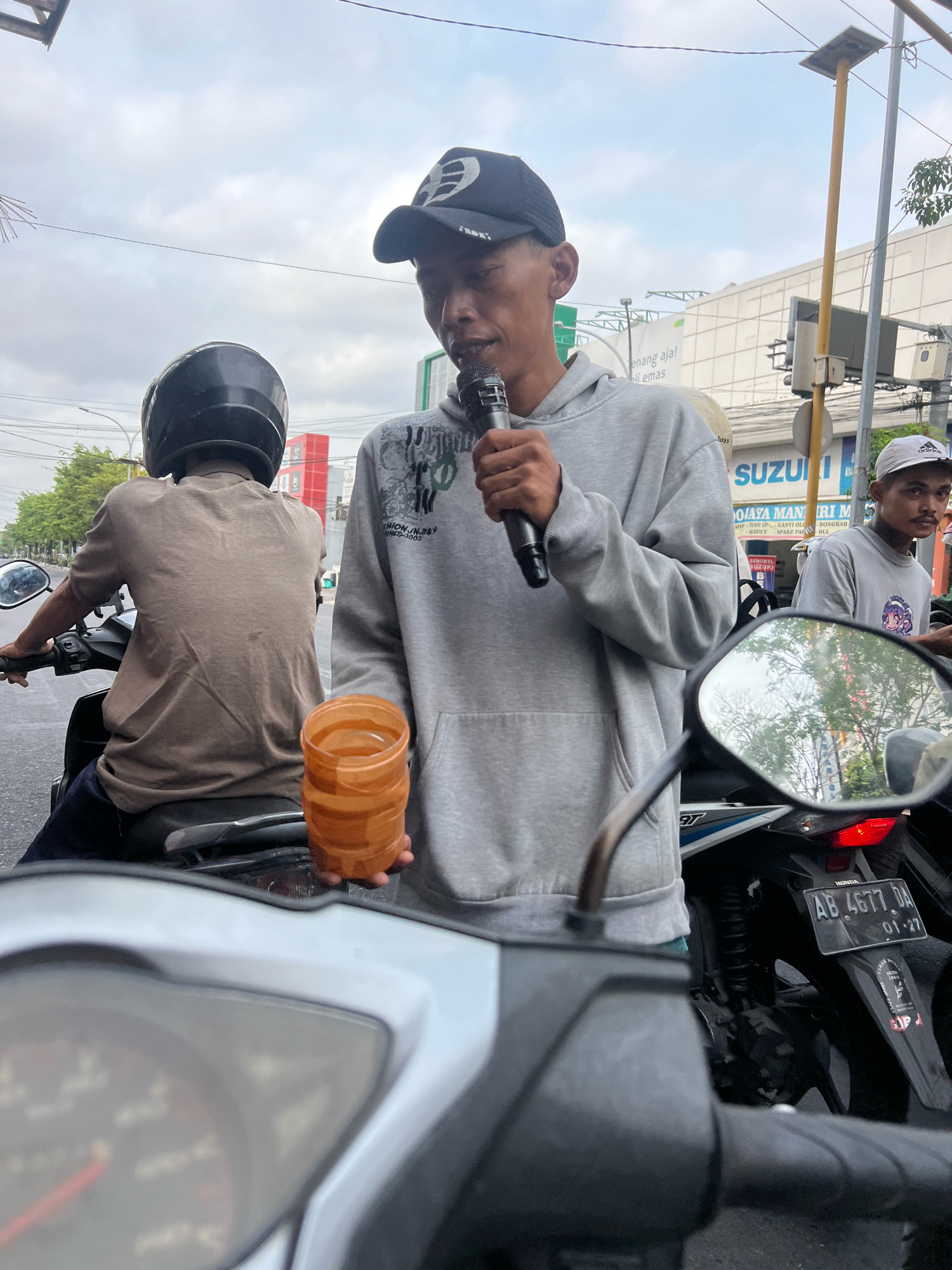 The image depicts a street scene in Yogyakarta, Indonesia, on a cloudy day. In the foreground, the handlebars and dashboard of a motorcycle are visible. To the right, a young man wearing a grey hoodie and a black baseball cap is holding a microphone in his right hand and a stack of orange plastic cups in his left. He appears to be speaking into the microphone. Behind him, another person on a motorcycle, wearing a brown shirt and a black helmet, is visible from the back. Further in the background, another young man wearing a white cap and a grey t-shirt is seen. A building with the "SUZUKI" logo is prominent in the background. A motorcycle license plate with "AB 4677 DA 01-27" is clearly visible. The overall atmosphere suggests a public street, possibly near a commercial area, with traffic present.