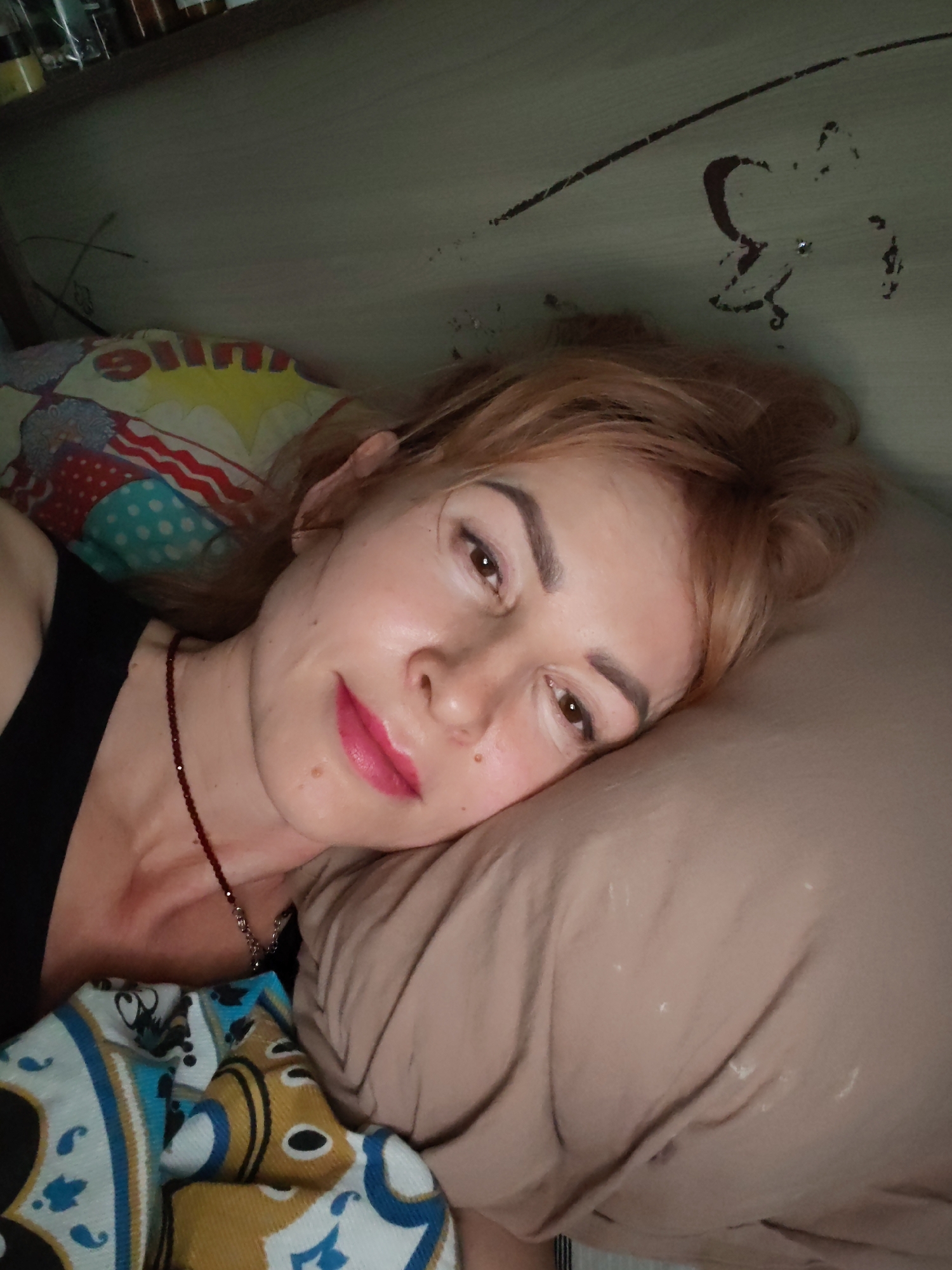 A woman with fair skin and red-blonde hair is lying on a beige pillow in what appears to be a bed. She is wearing a black tank top and a red beaded necklace. The bed is covered with a multi-colored patterned blanket. The wall behind the bed is painted a light color and has some markings or damage. A shelf with bottles is visible above the head of the bed. There is no specific indication of Chaikovsky, Russia in the image.