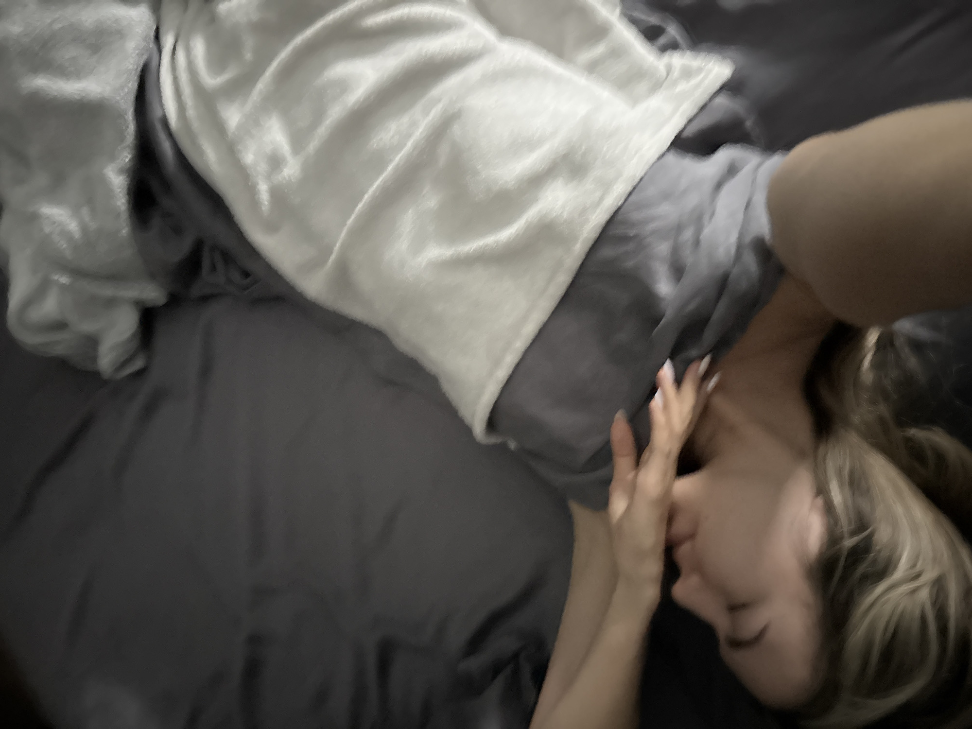 A close-up, overhead shot shows a person lying in bed, facing upwards with their eyes closed. Their right hand is raised and resting on their forehead, obscuring their eyes. Their hair, a shade of blonde, is spread out around their head. A white blanket is draped over their body, partially covering a grey blanket underneath. The scene is dimly lit, with the focus on the person and the textures of the bedding. The person appears to be resting or sleeping in a bedroom.