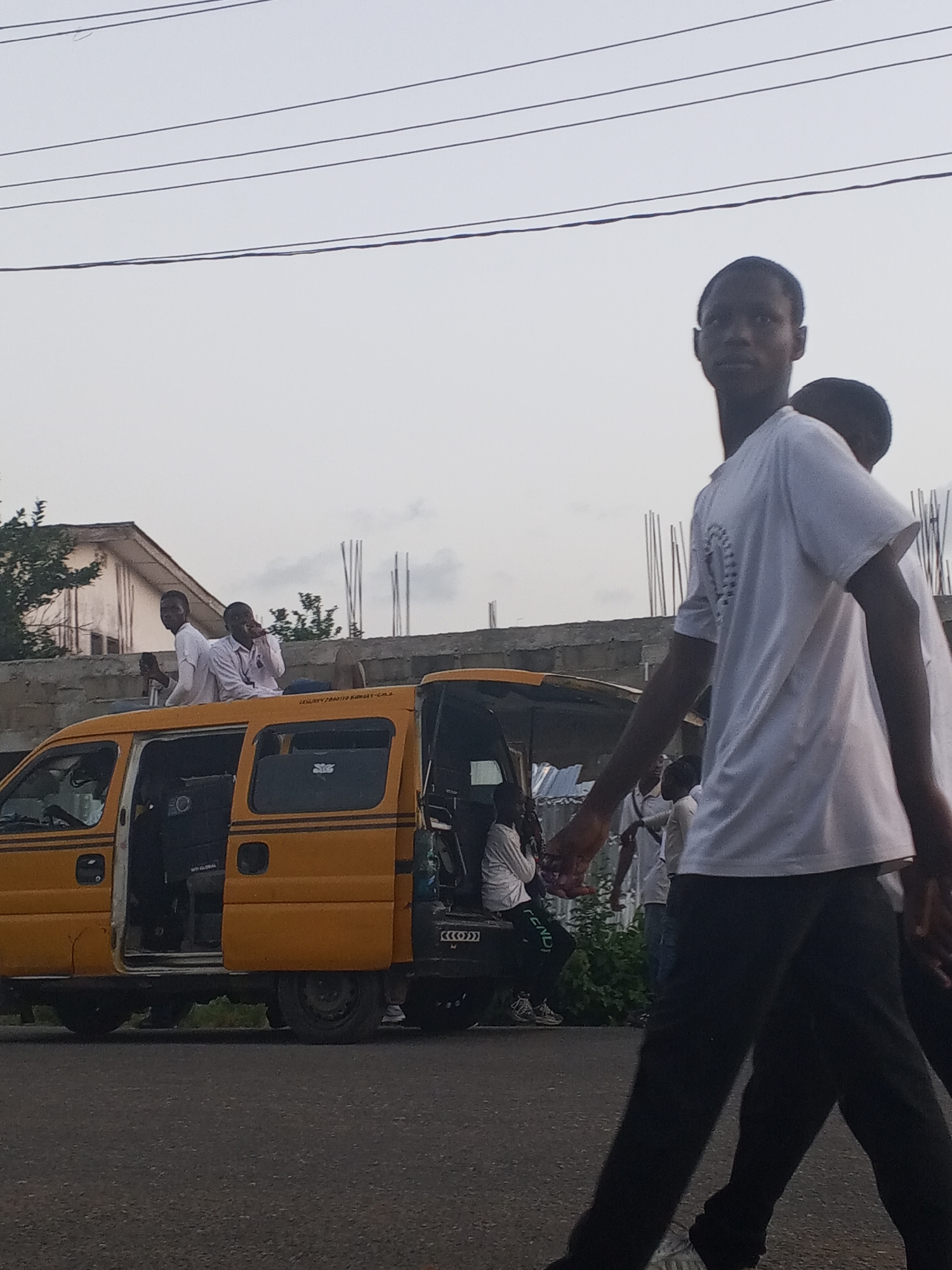 The photo shows a group of people, mainly young men, in Badagry, Nigeria. An open-doored yellow van with the license plate "CGC423XG" is the focal point. Two men are sitting on the roof of the van, and one is sitting inside the van. Other young men, wearing white t-shirts and dark pants, are walking in front of the van. Utility poles and lines are visible in the sky. A building under construction is in the background.