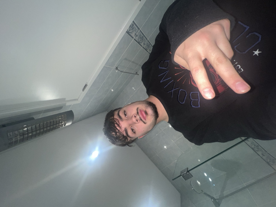 The image depicts a young adult male in an indoor setting located in the city of Vila Real, Portugal. The male has short, dark hair, a mustache, and a chin beard. He is wearing a dark, long-sleeved shirt with the word "BOXING" and a sunburst graphic visible on the front. His left hand is raised to chest level, displaying a V-sign gesture with the index and middle fingers extended. A white earbud is inserted into his right ear. The background features light grey tiled walls, with a horizontal band of patterned decorative tiles spanning the mid-section. A clear glass partition with a metallic hinge is visible on the left. The white ceiling contains a bright overhead light source and a rectangular, gridded ventilation or heating unit. A white cabinet or door is visible in the upper right portion of the frame. The male is looking directly at the camera.
