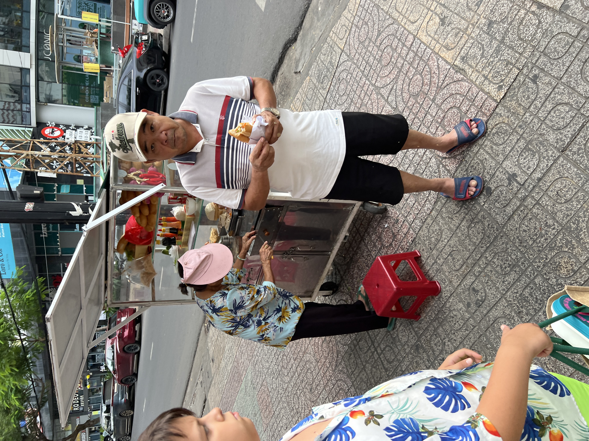 A man stands on a sidewalk in Nha Trang, Vietnam, holding a sandwich. He wears a white polo shirt with red and blue stripes, black shorts, a beige baseball cap with "Vietnam" written on it, and blue and red flip-flops. He is looking at the camera. To his left, a woman wearing a floral print shirt and a pink hat stands behind a food cart. The cart is made of metal and glass, displaying bread, bottles of sauce, and other food items. A red plastic stool is placed near the cart. In the background, cars are parked on the street and buildings line the sidewalk. The sidewalk is made of patterned concrete tiles. The scene appears to be a street food vendor stall.