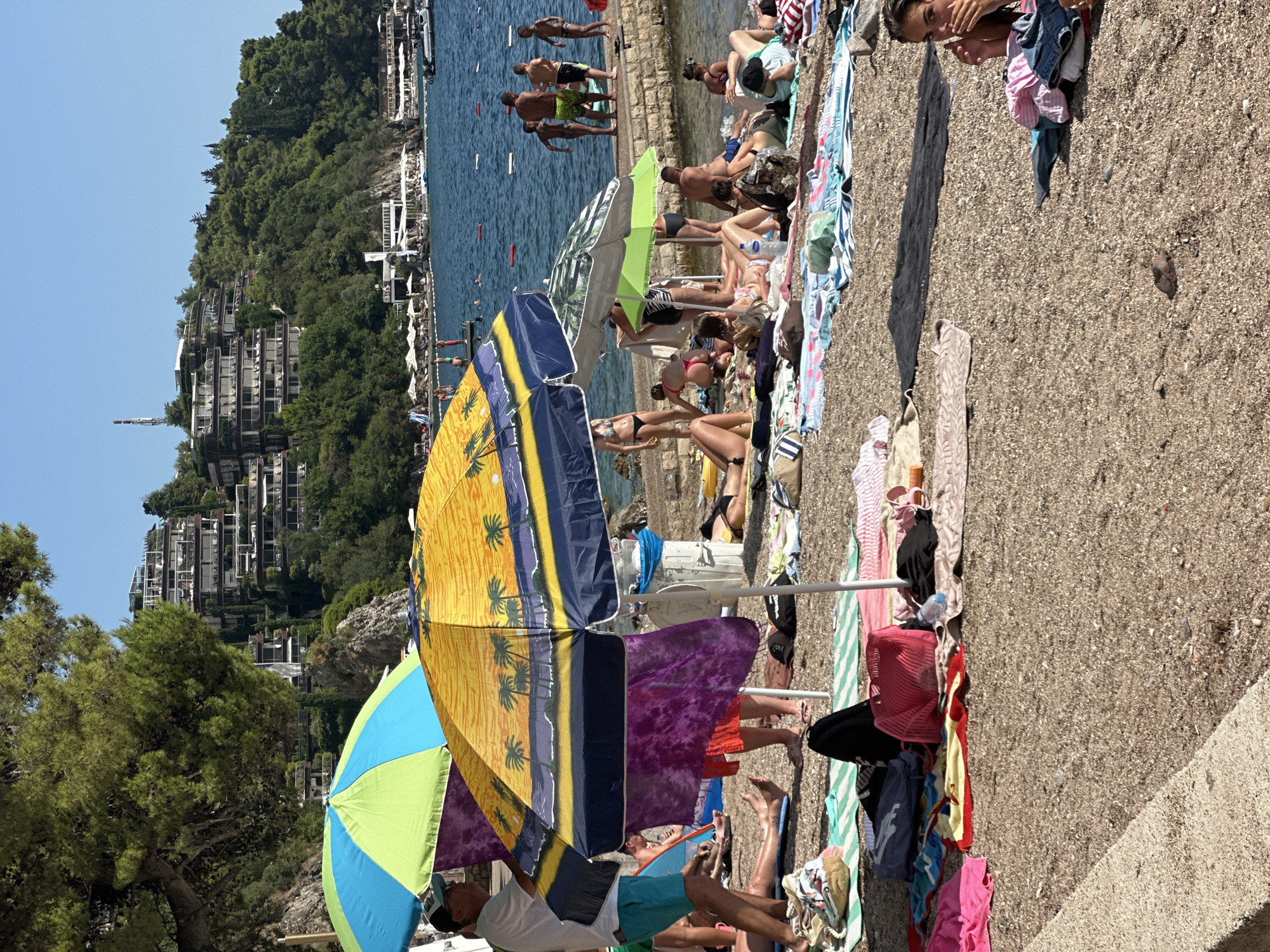 The image depicts a crowded beach scene in Budva, Montenegro. Several people are sunbathing and relaxing on the pebble beach, with towels and beach umbrellas scattered around. In the background, the turquoise water of the Adriatic Sea is visible, along with boats and a hillside covered in lush green trees and buildings. Two beach umbrellas, one with a vibrant yellow and blue pattern and another in green and blue, are prominently featured in the foreground, providing shade for beachgoers. People of various ages are present, some are lying on towels, others are sitting up and chatting. The weather appears to be sunny and warm, indicating it is likely midday during the summer season. The overall atmosphere is one of leisure and enjoyment associated with a typical beach holiday. There are no visible text elements in the image.