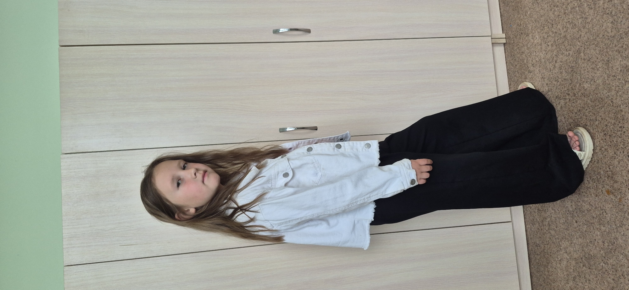 A young girl is lying down indoors in an unknown location in the city of Unknown. She has long, light brown hair and is wearing a white denim jacket over black pants and sandals. The background consists of a light-colored, wooden-looking cabinet or wardrobe. A portion of the room is carpeted with a brown material. A green wall is visible on the left side of the frame.