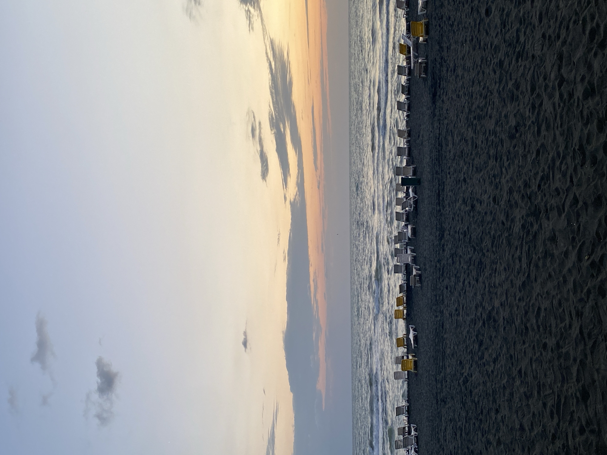 The image depicts a beach scene in Ureki, Georgia. The foreground consists of dark, textured sand, with ripples indicating the tide's movement. A long row of empty beach loungers is arranged on the sand, facing the sea. Some loungers are white, while a few are yellow. Beyond the sand, the sea stretches out, with gentle waves rolling towards the shore. The horizon is where the sea meets the sky, which is a gradient of soft colors, transitioning from pale blue at the top to warm orange and pink hues near the horizon, indicating either sunrise or sunset. Scattered clouds are visible in the sky. The overall atmosphere is serene and still, with no people present.