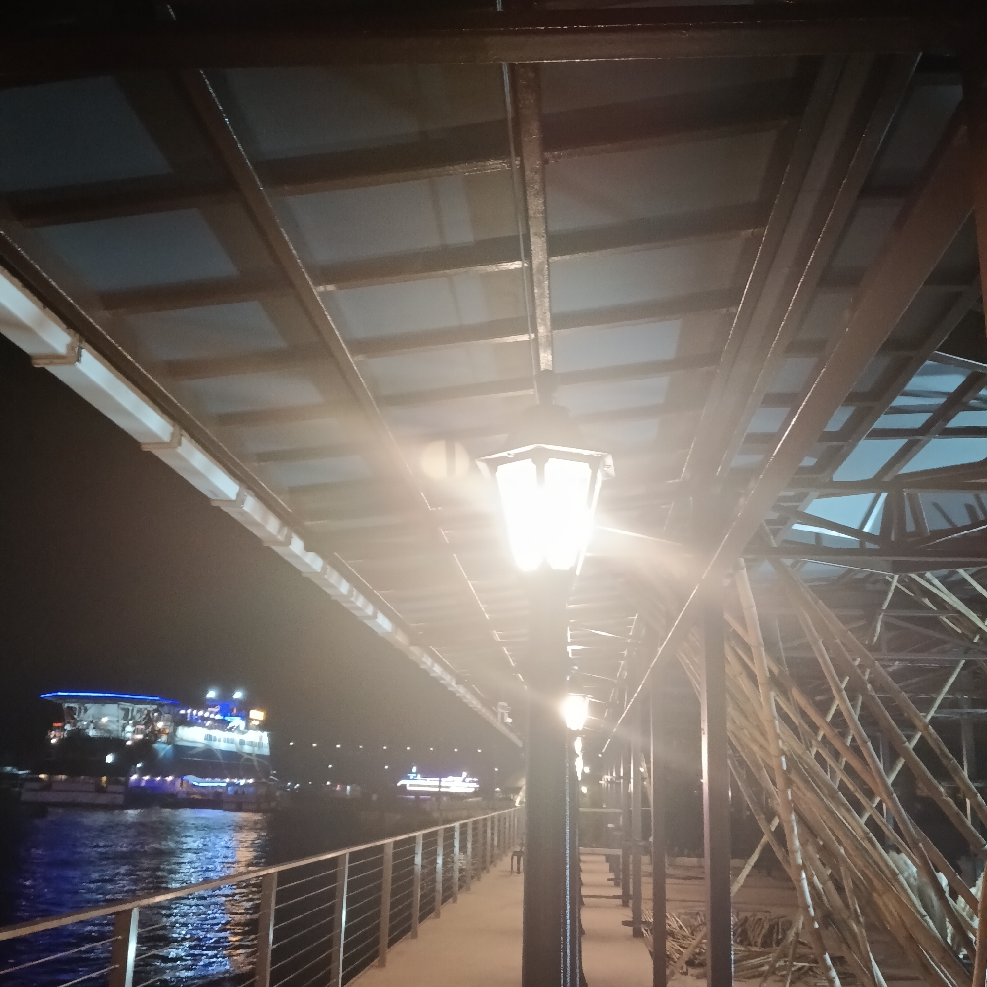 The image depicts a night scene on a pier or walkway in Panaji, India. In the background, a brightly lit boat floats on the water, reflecting the lights. The pier is lined with metal railings on the left side. A series of ornate, brightly lit lampposts illuminate the walkway. Above, a metal grid structure provides partial cover. To the right, bamboo scaffolding is visible, suggesting construction or renovation activity. No people are clearly visible in the image.