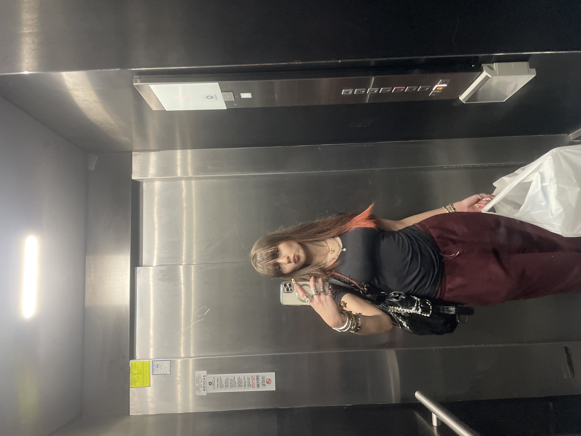 A woman is taking a selfie in an elevator. She is holding her phone in her left hand. She has long, brown hair with reddish highlights and is wearing a black top and reddish-brown pants. She has multiple rings and bracelets on her left hand. She is holding a white plastic bag in her right hand. The elevator appears to be made of stainless steel with black trim. There are elevator buttons and a light fixture visible above. There is no specific information in the image to indicate the location is Raanana, Israel.