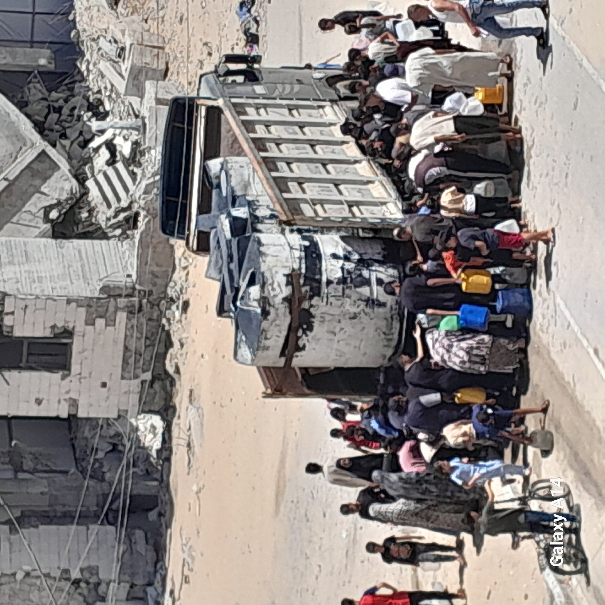 The image depicts a street scene in Deir el-Balah, Palestinian Territories, with a damaged building in the background. In the foreground, a large truck is parked, its bed filled with several large, cylindrical water tanks. A crowd of people, including men, women, and children, are gathered around the truck and in the street, many holding or collecting water from the tanks using various containers such as yellow jerry cans and blue barrels. Some individuals appear to be in the process of filling their containers, while others are observing or waiting. The ground is sandy and dusty. The overall atmosphere suggests a scene of resource distribution, likely water, in a community facing scarcity or in an area affected by damage.