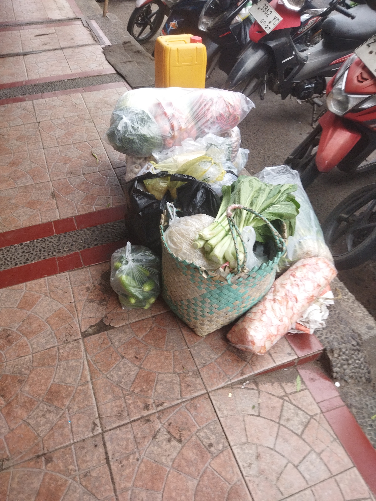 An overhead view shows an outdoor market scene on a tiled walkway in Banjar, Indonesia. Several motorcycles are parked behind a collection of goods, including a large yellow plastic container, bags of produce, a woven basket filled with green leafy vegetables, and a cylindrical object wrapped in pink and white plastic. The ground is covered in reddish-brown square tiles with a circular pattern. The overall impression is one of a bustling, everyday street market.
