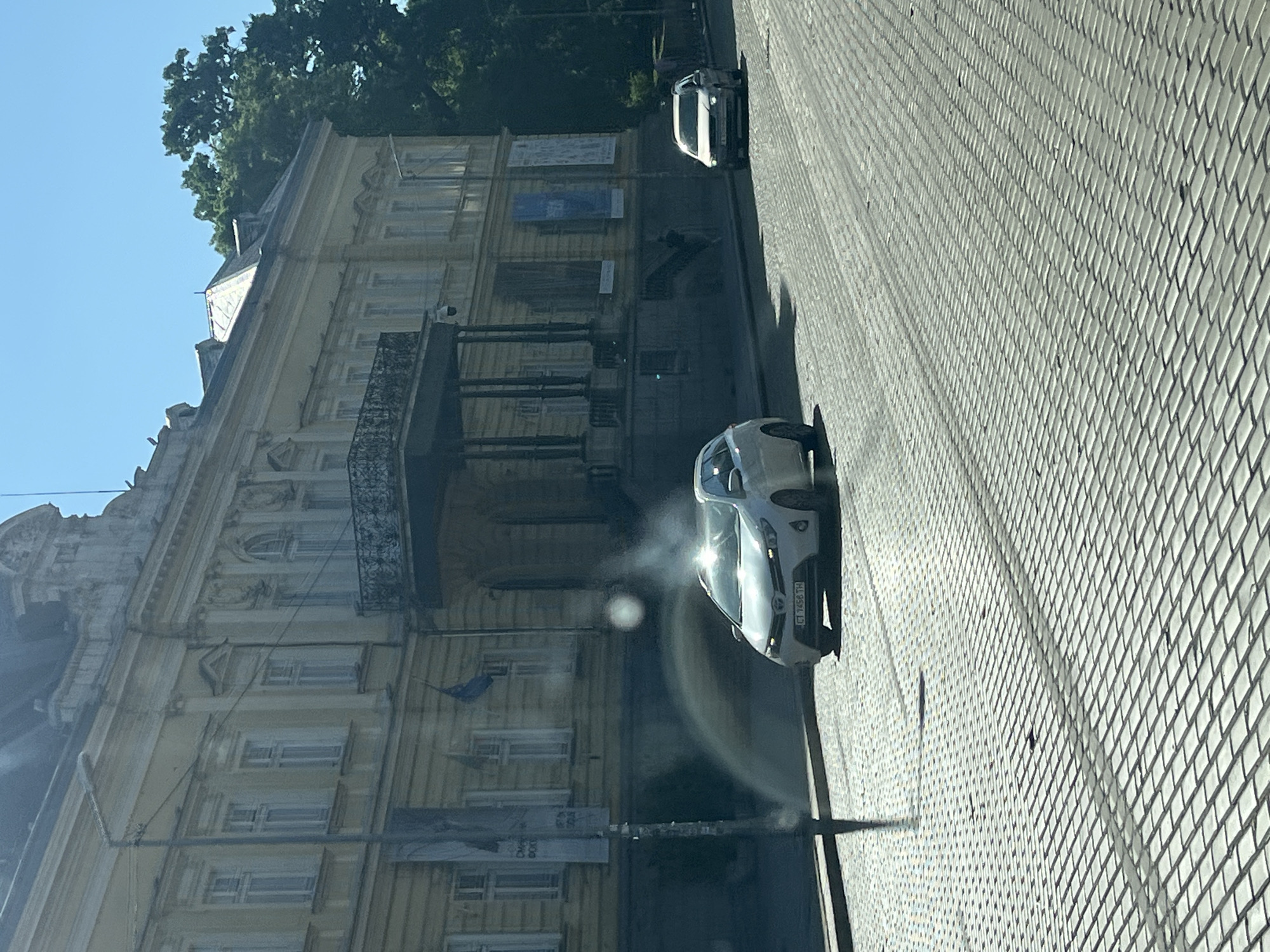 A white Toyota Auris is parked on a cobblestone street in front of a yellow, neoclassical building with ornate architectural details. The building has a balcony with wrought-iron railings and several tall windows. A lamppost stands on the left side of the image, and trees are visible in the background. The sky is clear and blue. The license plate of the car reads "CT 7456 TH".