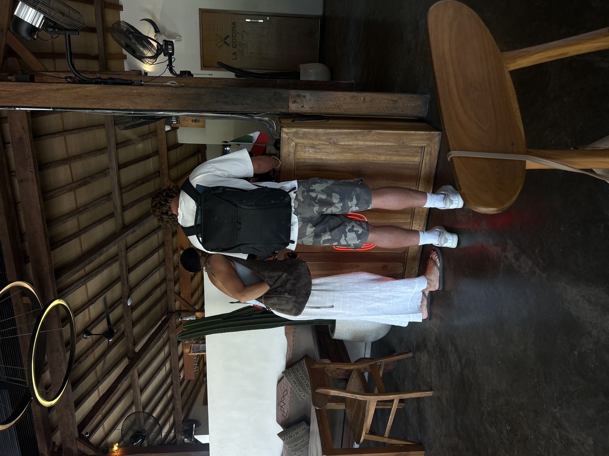 A man and a woman stand at a wooden counter in what appears to be a restaurant or reception area. The man, facing away from the camera, wears a white t-shirt, camouflage shorts, white socks, and white sneakers. He has a black backpack on. The woman, also facing away, wears a light-colored top and a long white skirt. She has a dark brown shoulder bag. Behind them is a wooden counter with some flags and what looks like a cactus plant. A door with "LA COCINA staff only" written on it is visible in the background. The ceiling has exposed wooden beams and a modern light fixture. There are also fans mounted on the wall and ceiling. In the foreground, a wooden chair is partially visible. The flooring is dark and appears to be concrete. The setting is likely a business or hospitality establishment in Kuta Selatan, Indonesia, given the presence of the Mexican flag and the overall aesthetic which is consistent with establishments in that region.