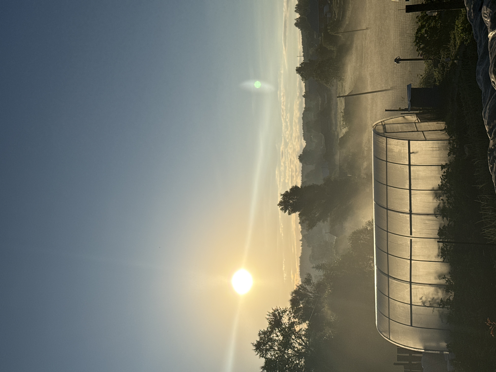This image depicts a greenhouse in a rural setting at sunrise. The sun, a bright orb with lens flare, is positioned in the upper left quadrant of the sky, casting a warm, golden light. A faint mist hangs in the air, creating a hazy atmosphere that softens the landscape. The greenhouse, a translucent, arched structure, occupies the lower left of the frame, illuminated from within and behind by the rising sun. Silhouetted against the misty background are trees and the distant shapes of buildings, suggesting a small village or settlement. The sky transitions from a pale yellow near the horizon to a clear, light blue at the top. There are no people visible in the image.