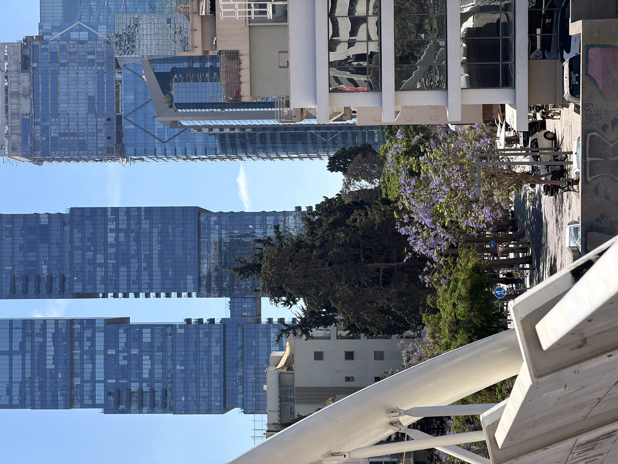 This image captures a street scene in Tel Aviv, Israel, with a blend of modern architecture and natural elements.
In the background are tall glass skyscrapers, reflecting the blue sky. In the midground, a tree is in full bloom with purple flowers. Below the tree there is a sidewalk with parked vehicles, pedestrians, and bicycle signs. In the foreground is a white metal structure, possibly part of a bridge.