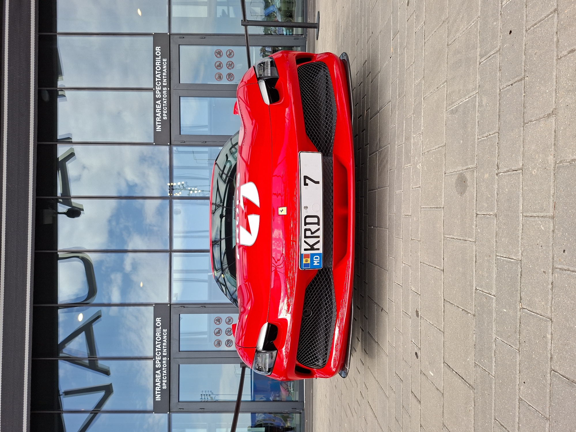 A low-angle, eye-level, full front-facing medium shot captures a bright red Ferrari sports car parked on a paved area outside a modern building. The building features large glass doors with signage in Romanian and English that reads "INTRAREA SPECTATORILOR" and "SPECTATORS ENTRANCE." Reflected in the glass are clouds and the sky, as well as what appears to be a large, dark logo or text. The car's license plate is visible and reads "MD KRD 7." The car has a black grille and a white graphic on its hood, which appears to be a stylized "2" or "Z" with a circle. The paved area is made of grey rectangular bricks. The overall scene is well-lit, suggesting daylight. No people are clearly visible, though the context of "Spectators Entrance" suggests a public venue. The specific location in Stăuceni, Moldova, is indicated by the license plate's "MD" designation.