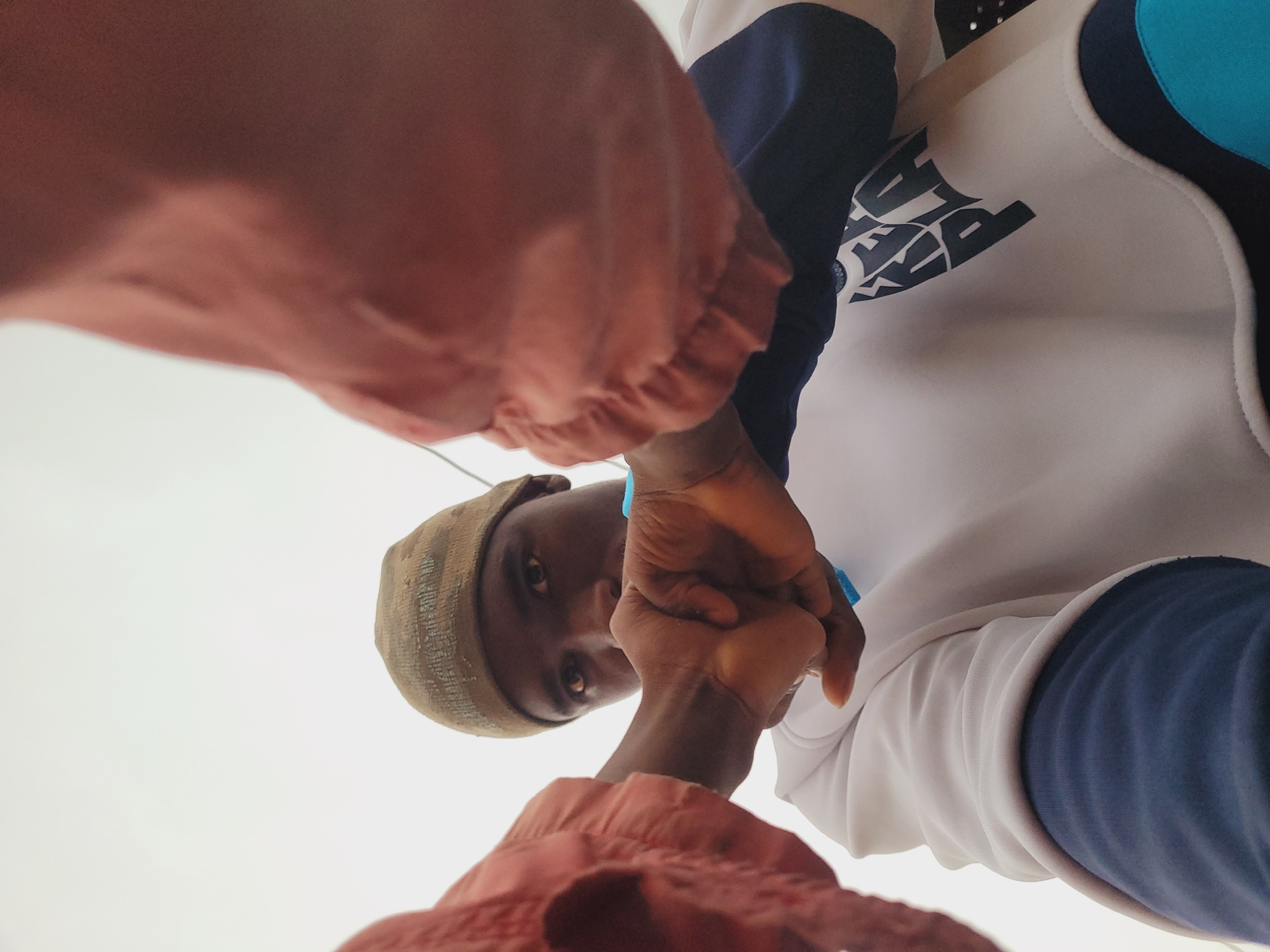 In Bukuru, Nigeria, a close-up, low-angle shot captures a person wearing a beanie and a white and blue sweatshirt. The person's face is partially obscured as they look directly at the camera, holding up a fist. Two pieces of rust-colored fabric are visible, one on the left and one on the right, framing the central subject. The background is a plain, bright sky.