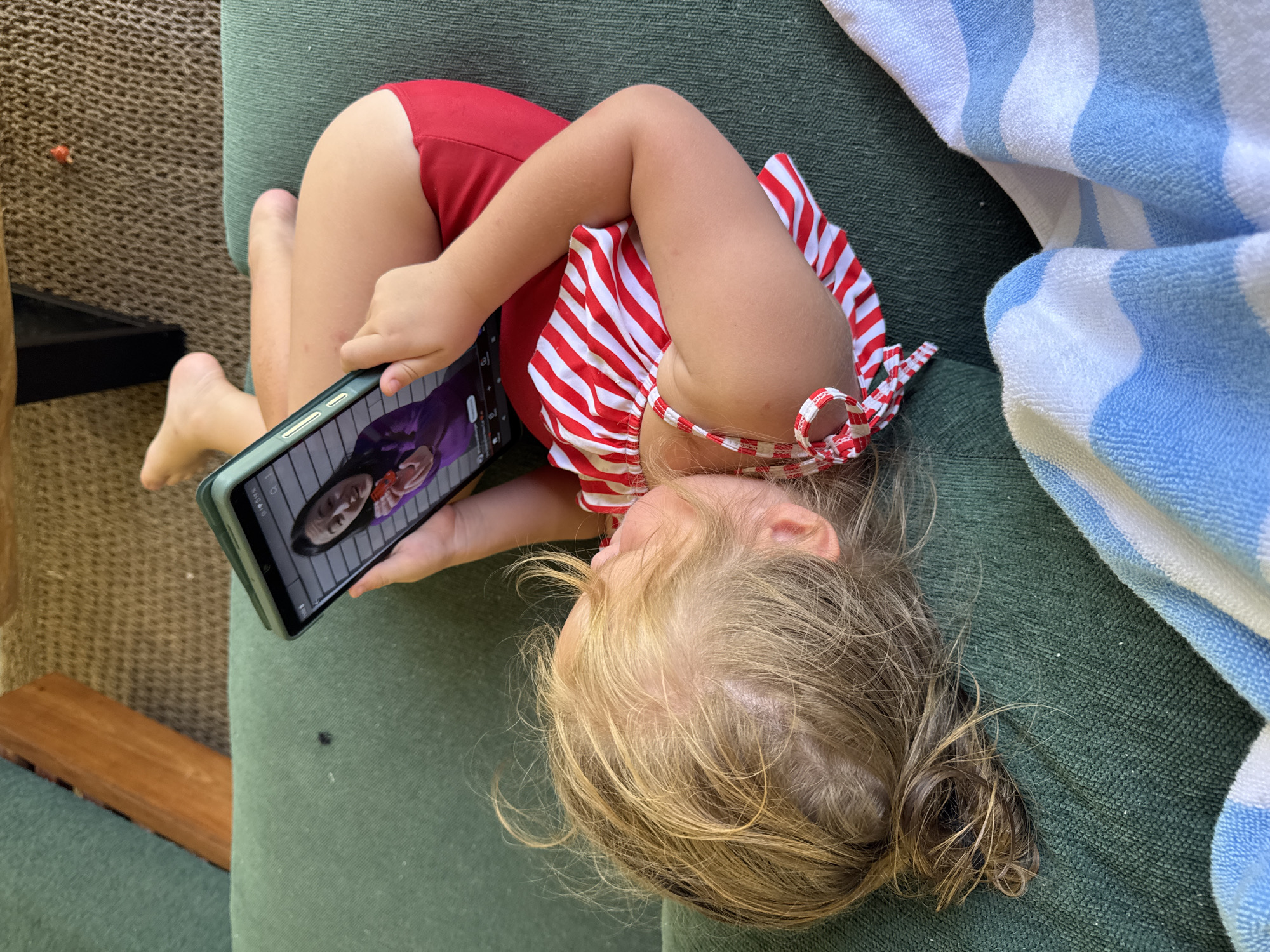 An overhead shot captures a young child, likely a toddler, lying on their side on what appears to be green outdoor lounge furniture, possibly a sunbed or cushion. The child has fair skin and light blonde hair, slightly disheveled. They are wearing a red swimsuit with a red and white striped ruffled top, visible on their upper back and shoulder, and solid red bottoms.
The child is intently focused on a smartphone held horizontally in both hands. The phone screen displays a person's face, appearing to be a video call or a video, featuring an individual with dark hair wearing a light-colored top, possibly against a purple or dark background. Small interface icons, such as battery and signal strength indicators, are visible at the top of the phone screen, but no legible text is discernable.
Beside the child, a blue and white striped towel is crumpled on the green surface. In the background, to the left, another textured surface, possibly a woven rug or part of a different piece of furniture, and a wooden slat are visible. The lighting suggests it is daytime, indicative of warm weather suitable for swimwear. The scene conveys a relaxed moment, likely taking place outdoors in Kuta, Indonesia.