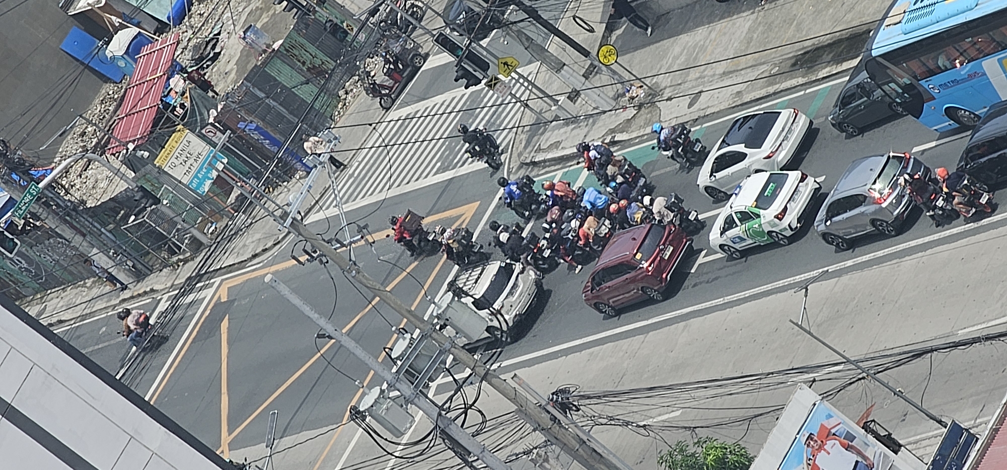 This is an aerial, vertical, full shot of a busy intersection in Makati, Philippines, on a sunny day. The scene is dominated by a dense traffic of motorcycles, cars, and a bus. The road is paved and marked with yellow lines. A pedestrian crossing is visible. Utility poles with a complex network of wires are scattered throughout the frame. On the right side of the image, there are makeshift structures, possibly informal settlements, with corrugated metal roofs and tarps. A billboard with a picture of a man is visible on the left side. The overall impression is one of urban congestion and informal development coexisting with modern infrastructure.