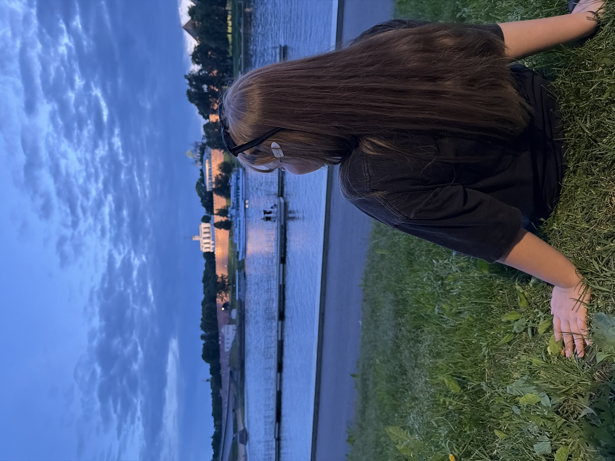 A person sits on a grassy embankment facing a river at dusk. In the background, across the river, is a fortress wall with buildings and a prominent white structure with domes and towers. A bridge spans the river to the left. A boat is docked on the river in the middle ground. The sky is cloudy and dark blue. The person is wearing a black t-shirt and has long, brown hair. They are wearing sunglasses on their head and have small white clips in their hair. Their left hand is resting on the grass. The scene appears to be the Veliky Novgorod Kremlin on the banks of the Volkhov River in Veliky Novgorod, Russia.