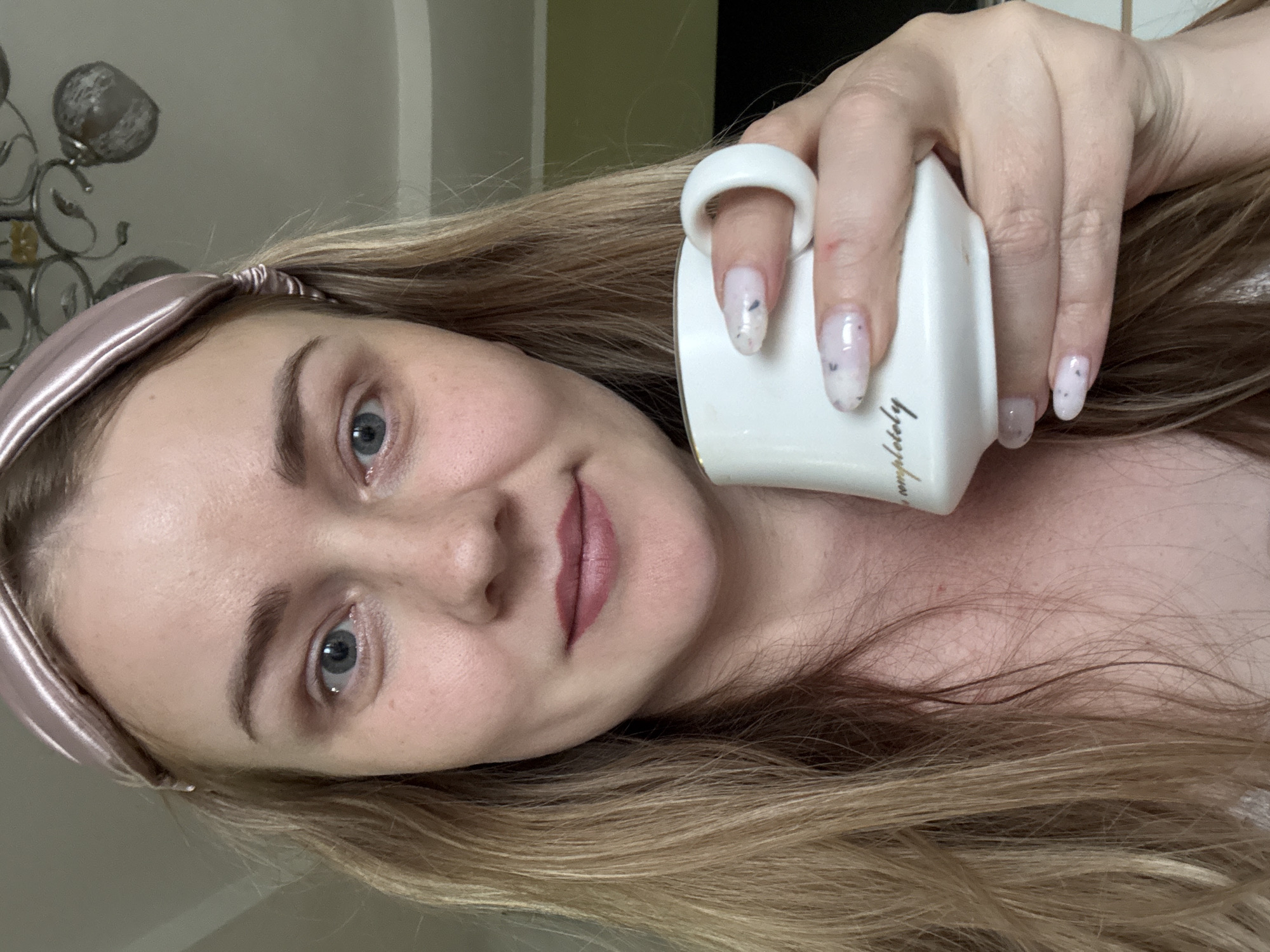 A woman is holding a white mug with the word "Completely" printed on its side. She has blonde hair, blue eyes, and is wearing a light pink headband. The background appears to be an interior space with a decorative lamp visible in the upper left corner. The city of Chaikovsky, Russia, cannot be determined from the image.