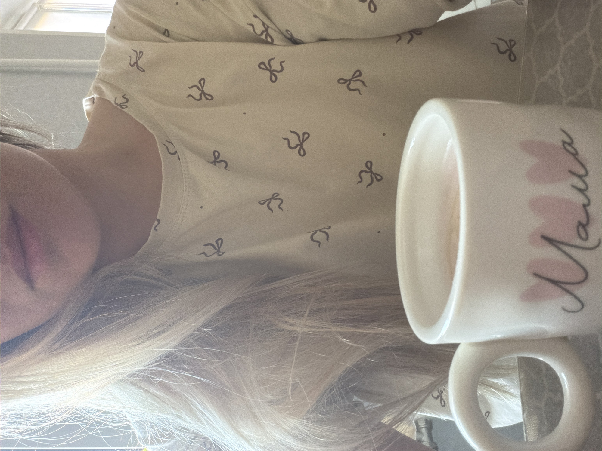 A low-angle, close-up photograph shows a fair-skinned person with long blonde hair next to a white mug with the word "Mama" in cursive with pink heart accents. The person is wearing a light-colored, long-sleeved shirt patterned with small black bows. Part of a window with white trim is visible in the background. The image gives no indication of a location in the city of Votkinsk, Russia.