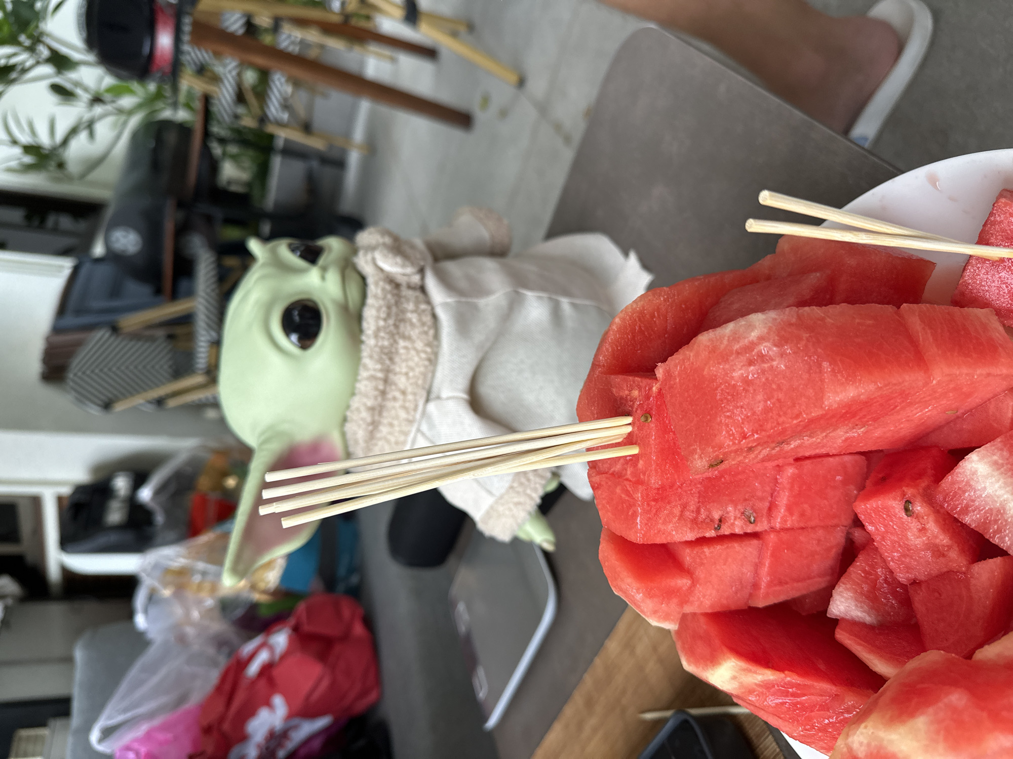 A full shot shows a table with a bowl of sliced watermelon and a Grogu doll. The watermelon is in a white bowl in the foreground, and several bamboo skewers are inserted into the fruit. The Grogu doll, a character from Star Wars, is in the middle ground, wearing a tan robe and scarf. In the background, there are chairs, tables, plants, and various bags. A person's feet are visible in the lower right corner, wearing white slides. The image appears to be taken outdoors, possibly on a patio or balcony.