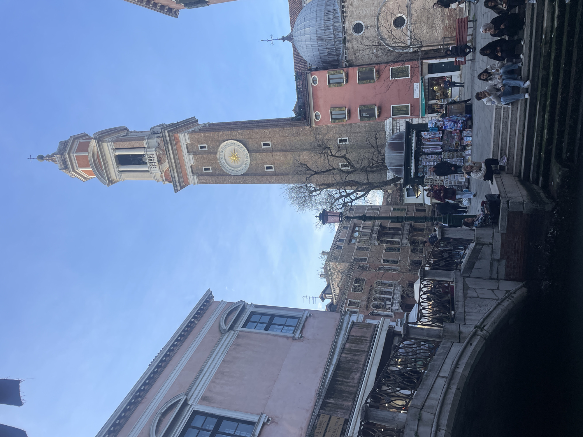 The image shows a street scene in Venice, Italy. A tall brick campanile with a large clock is visible on the left. A domed building is to the right of the campanile. A bridge is in the foreground, with people sitting on steps leading up to the square. There is a souvenir stand next to the steps. Several buildings in the background have multiple windows and traditional Venetian architecture. The sky is a clear blue.