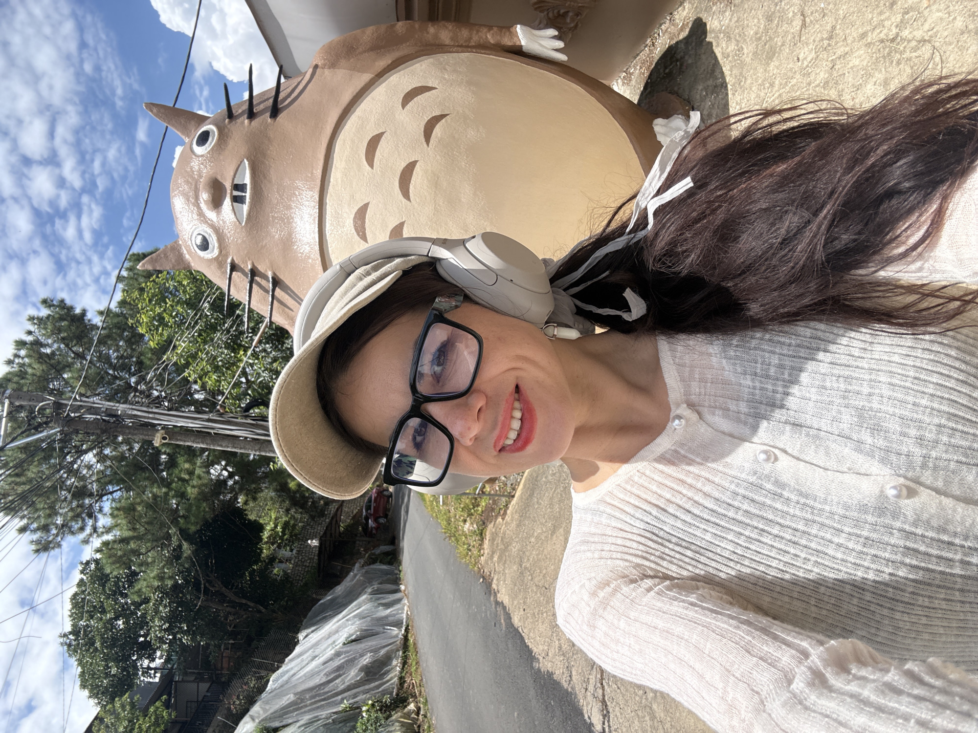 The image shows a woman taking a selfie outdoors in Da Lat, Vietnam, with a large statue of the character Totoro from the Studio Ghibli film "My Neighbor Totoro" in the background. The woman is wearing a beige baseball cap, large black-rimmed glasses, and a light-colored, long-sleeved, button-up cardigan adorned with pearls. She has on headphones and her dark hair is partially pulled back, with white ribbons tied into it. She is smiling and looking at the camera.
The Totoro statue is brown and beige, with large eyes, a wide mouth showing two front teeth, and whiskers. It is positioned behind the woman, taking up a significant portion of the frame. The background includes a paved road that curves uphill to the left, with a sloped area covered in white plastic sheeting on the left side of the road, possibly a greenhouse or agricultural covering. Trees and vegetation are visible on the hillside. A utility pole with wires stands to the right of the woman and in front of the Totoro statue. The ground in the foreground is concrete. The sky is blue with scattered white clouds. A red car is partially visible parked on the road in the distance.