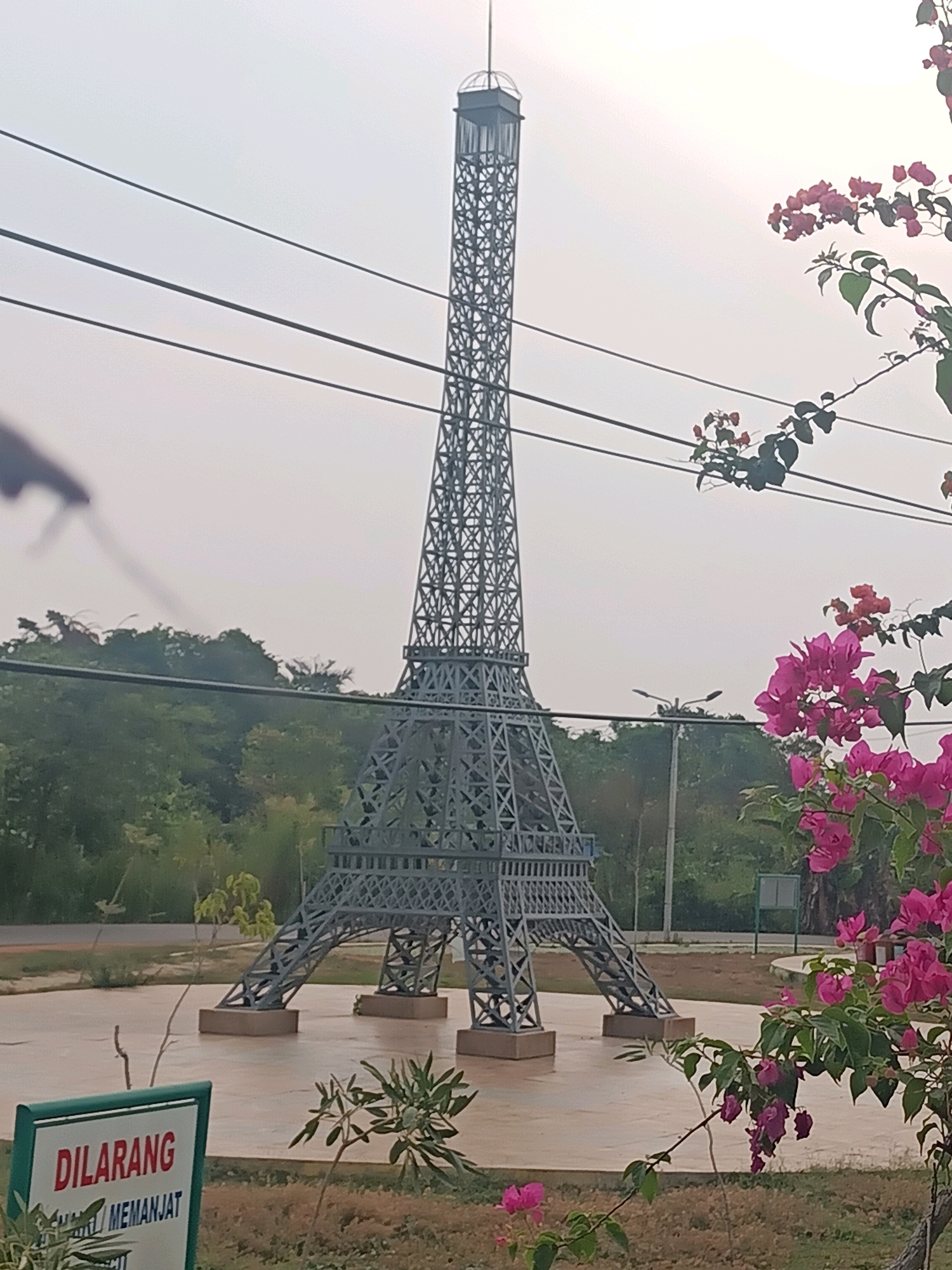 This image depicts a miniature Eiffel Tower replica, located in the city of Bagus, Indonesia. The replica is grey metal, with intricate latticework. It stands on a circular concrete base in an open, grassy area. In the foreground, a sign with green trim displays text in red and black. Bright pink bougainvillea flowers are visible in the right side of the frame, framing the tower. The background consists of a dense green tree line and a hazy, pale sky. Power lines are strung across the upper portion of the image.