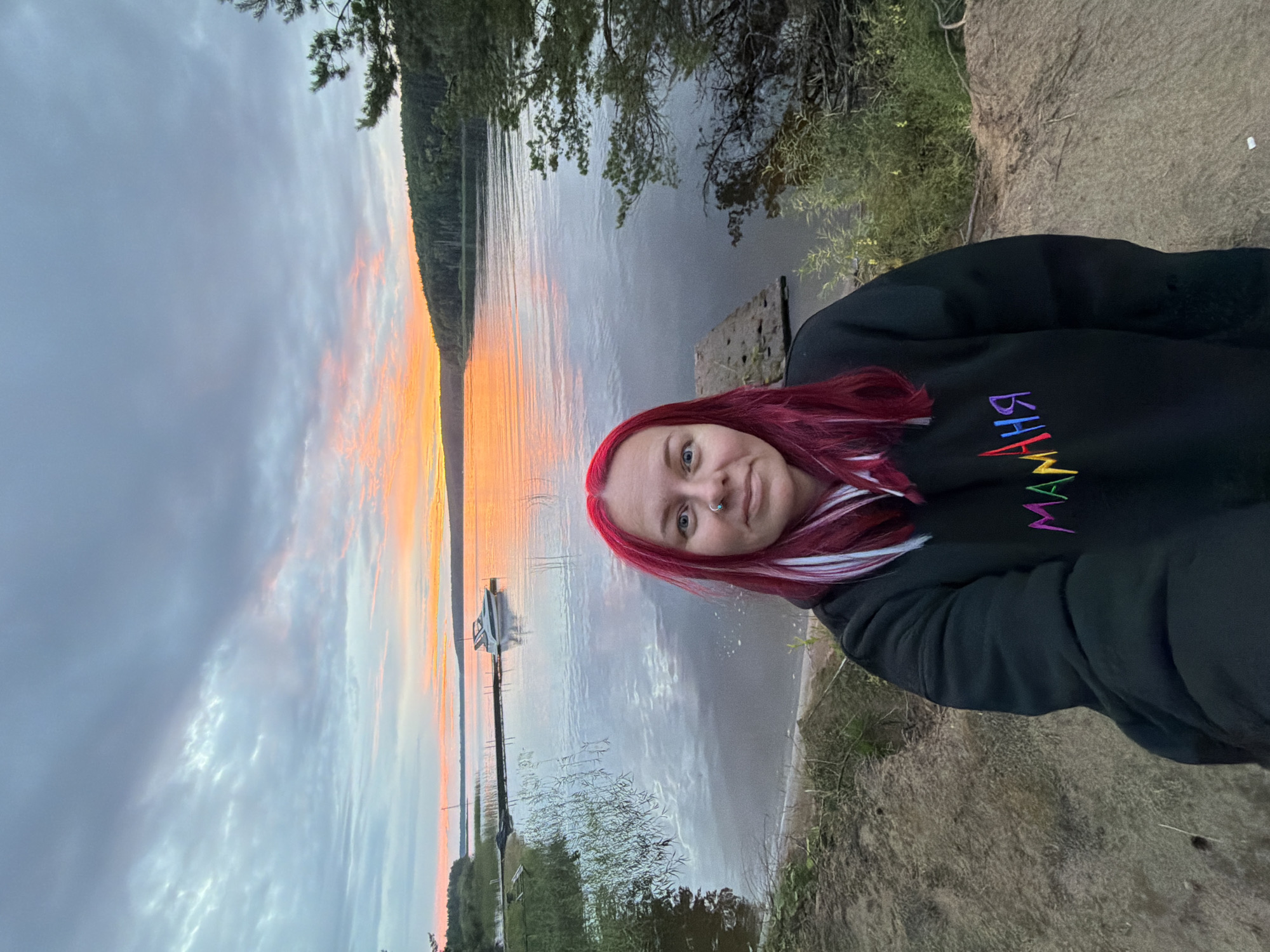 A woman with vibrant red and white dyed hair is taking a selfie outdoors. She is wearing a black hoodie with the word "BHANM" embroidered on it in rainbow lettering. Her expression is neutral and she is looking towards the camera.
The setting is a natural landscape with a body of water, likely a lake or river, and a treeline along the far bank. The water reflects the sky, which is filled with soft clouds tinged with the warm hues of either sunrise or sunset, suggesting it is early morning or late evening. A small boat is docked at a pier extending into the water. The ground in the foreground is rough and earthy, with some sparse vegetation.
The overall mood is serene and tranquil, with the natural beauty of the scene being a prominent element. The time of day is indicated by the colors in the sky.