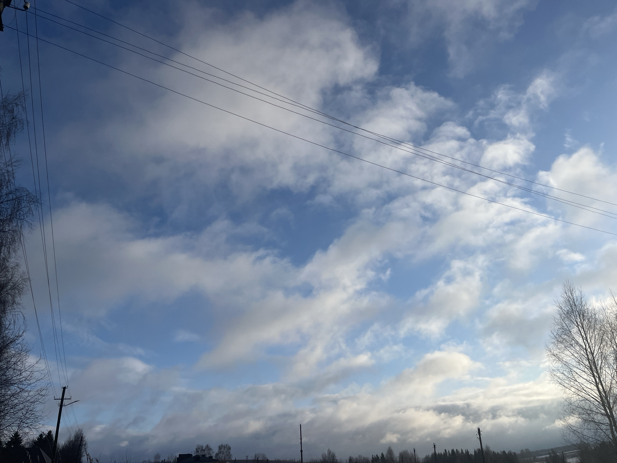 The image shows a winter sky in Павловка, Russia, filled with puffy white and grey clouds interspersed with patches of blue. Bare trees are visible along the horizon, along with telephone poles and power lines. The overall impression is of a cold, overcast day.