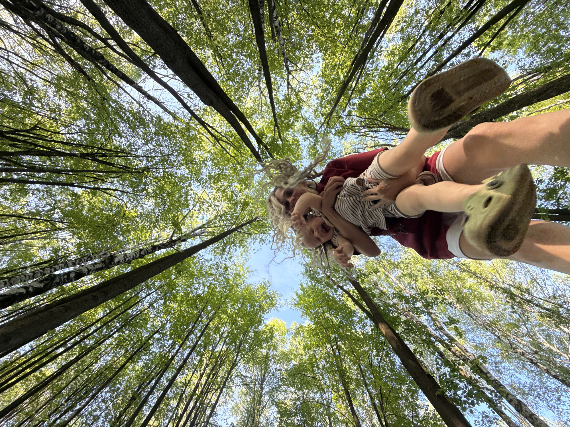 A low-angle shot looking up at a person holding a child overhead in a forest. The camera is aimed upward, capturing the canopy of tall, thin trees with green leaves against a blue sky. The person holding the child appears to be an adult with blond dreadlocks, wearing a red shirt and white shorts. The child is wearing a striped shirt and holding onto the adult. The trees are dense, filling the frame, with only small patches of sky visible. The scene is filled with natural light, creating a sense of being immersed in the woods. The location is presumed to be a forest near Rusanovo, Russia based on the prompt's instructions.