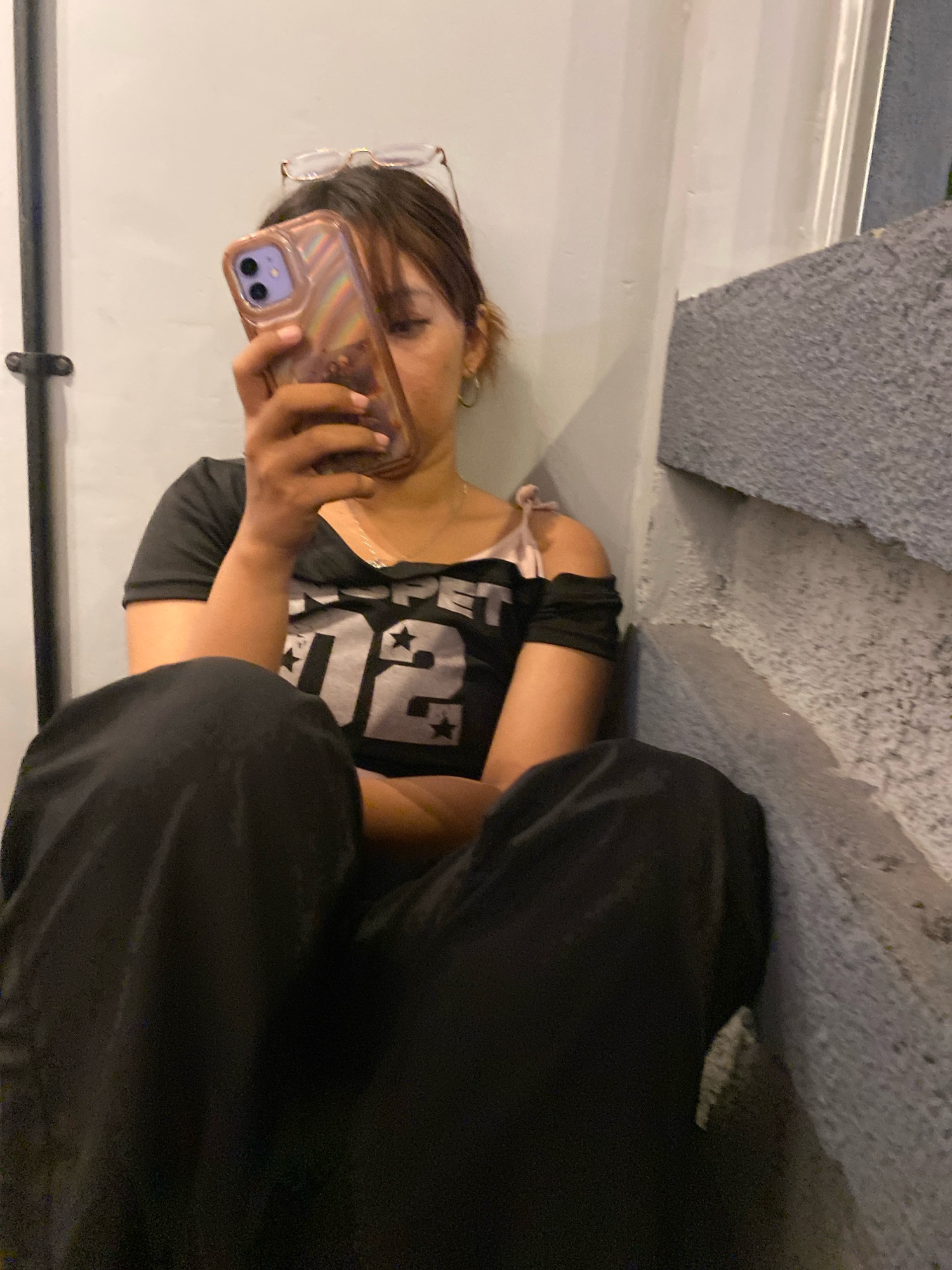 A young woman with brown hair and glasses perched on her head sits on the ground, leaning against a concrete wall. She is wearing a black t-shirt with white lettering and black pants. She is holding a smartphone in her hands, looking down at the screen. The background is a textured gray wall, suggesting an urban environment. The lighting is dim, creating a somewhat shadowed atmosphere. The setting appears to be an outdoor area, possibly a street or alleyway, in Klurak, Indonesia.