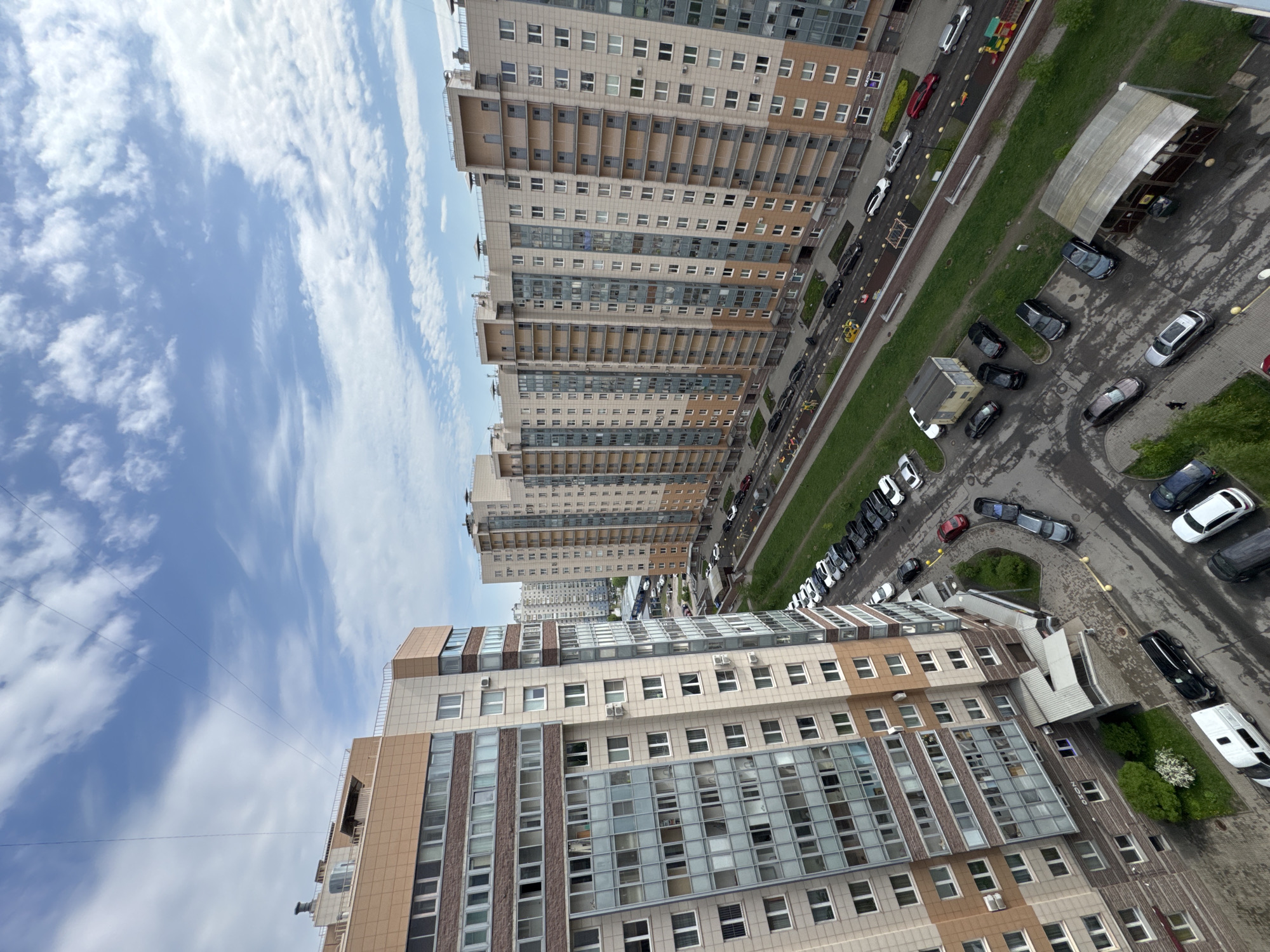 The image shows an urban scene in Vsevolozhsk, Russia, featuring a view from an elevated perspective, potentially from a high-rise building. Multiple tall, modern apartment buildings dominate the scene with beige, brown, and glass facades. The sky is blue with streaky clouds. Below, a road runs parallel to the buildings, with several parked cars and a few moving vehicles. A playground is visible adjacent to the road. Further below and in front of the nearest building is a parking area with several cars. There is evidence of wet pavement or rain. The image appears to be taken during the daytime.