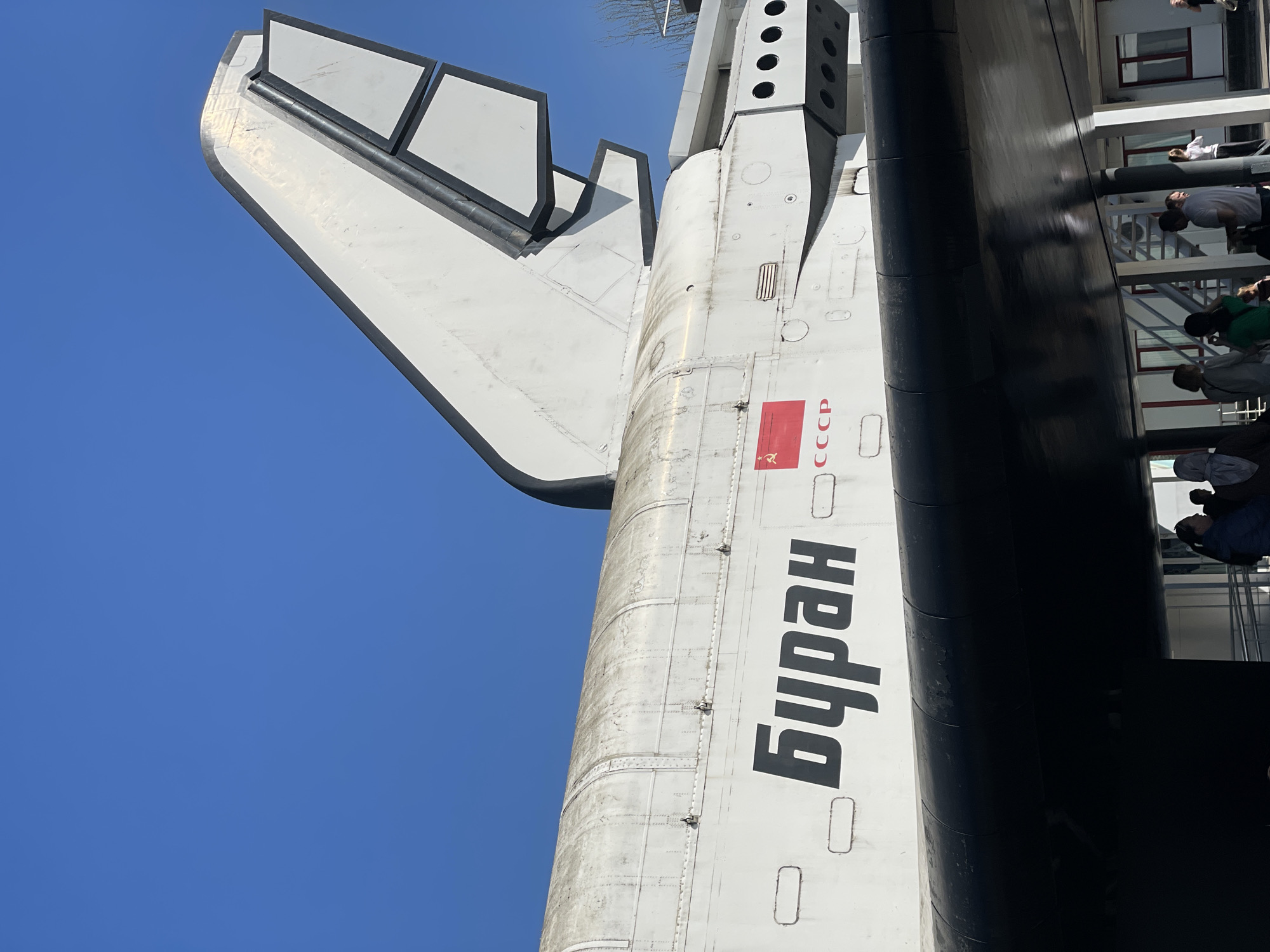 The image shows the tail of the Buran spacecraft, situated in Khimki, Russia. The spacecraft has "Buran" written vertically in black Cyrillic letters on its white fuselage, accompanied by the Soviet Union flag and "CCCP" lettering. The background features a clear blue sky, a building structure with windows, and a viewing platform with people. Some of the people are interacting with the spacecraft or the viewing platform. The spacecraft's tail fin structure is visible, with rectangular outlines. The overall environment appears to be an outdoor exhibit or museum setting.