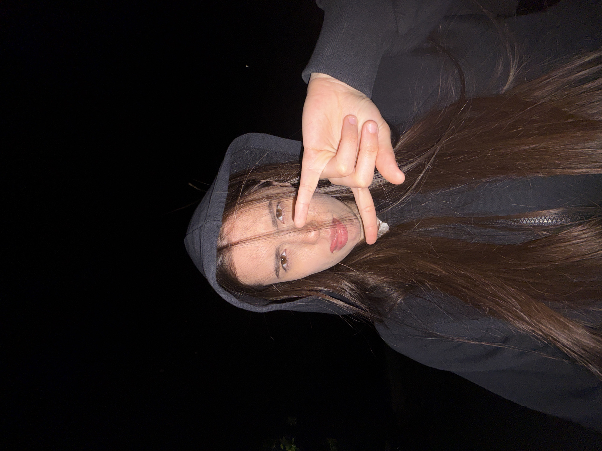 A young woman with long, dark brown hair is wearing a dark hooded sweatshirt and making a hand gesture with her fingers. The background is dark, with little to no discernible detail. It is night. The location is presumed to be Chaikovsky, Russia.
