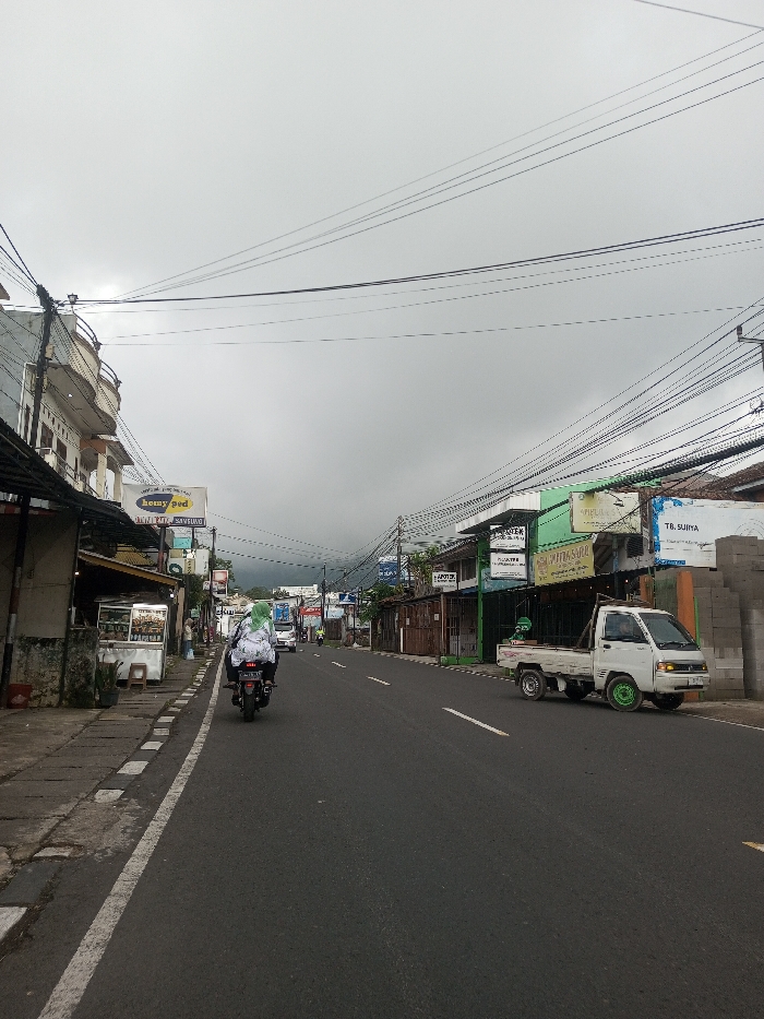 The image presents an eye-level view of a bustling street in what appears to be a town or village under a heavily overcast, grey sky. The asphalt road, marked with a solid white line on the left shoulder and a dashed white line in the center, stretches into the distance.
On the left side of the street, a person riding a black motorcycle away from the camera is visible in the middle ground. The rider is wearing a white and black patterned jacket and a distinctive bright green head covering. Adjacent to the road is a paved sidewalk, along which a row of buildings stands. In the foreground, there's a multi-story building with balconies, and further down, various commercial establishments and stalls are visible. One prominent sign on the left reads "heny pod," and a larger sign above it clearly displays "SAMSUNG." Another sign below "heny pod" shows partial text like "Dapur..." and "Coffee." A street vendor stall with food items is also visible on the left.
On the right side of the street, a small white pickup truck (ute) is parked, facing away from the camera. Its front two wheels are fitted with striking bright green hubcaps or rims. Behind the truck, a series of single-story commercial buildings line the road, some featuring green facades or awnings. A pile of grey concrete blocks is stacked beside one of these buildings on the far right. Several signs are visible on this side, including "APOTEK" (Pharmacy) and "TB. SUJIYA."
A dense network of black power lines and communication cables crisscrosses high above the street, a prominent feature of the overhead space. The overall lighting is soft and diffused due to the overcast conditions. In the far distance, beyond the buildings, the faint silhouettes of hills or mountains are discernible through the atmospheric haze. The scene depicts an active, everyday environment with various vehicles and open businesses, characteristic of urban or semi-urban areas.