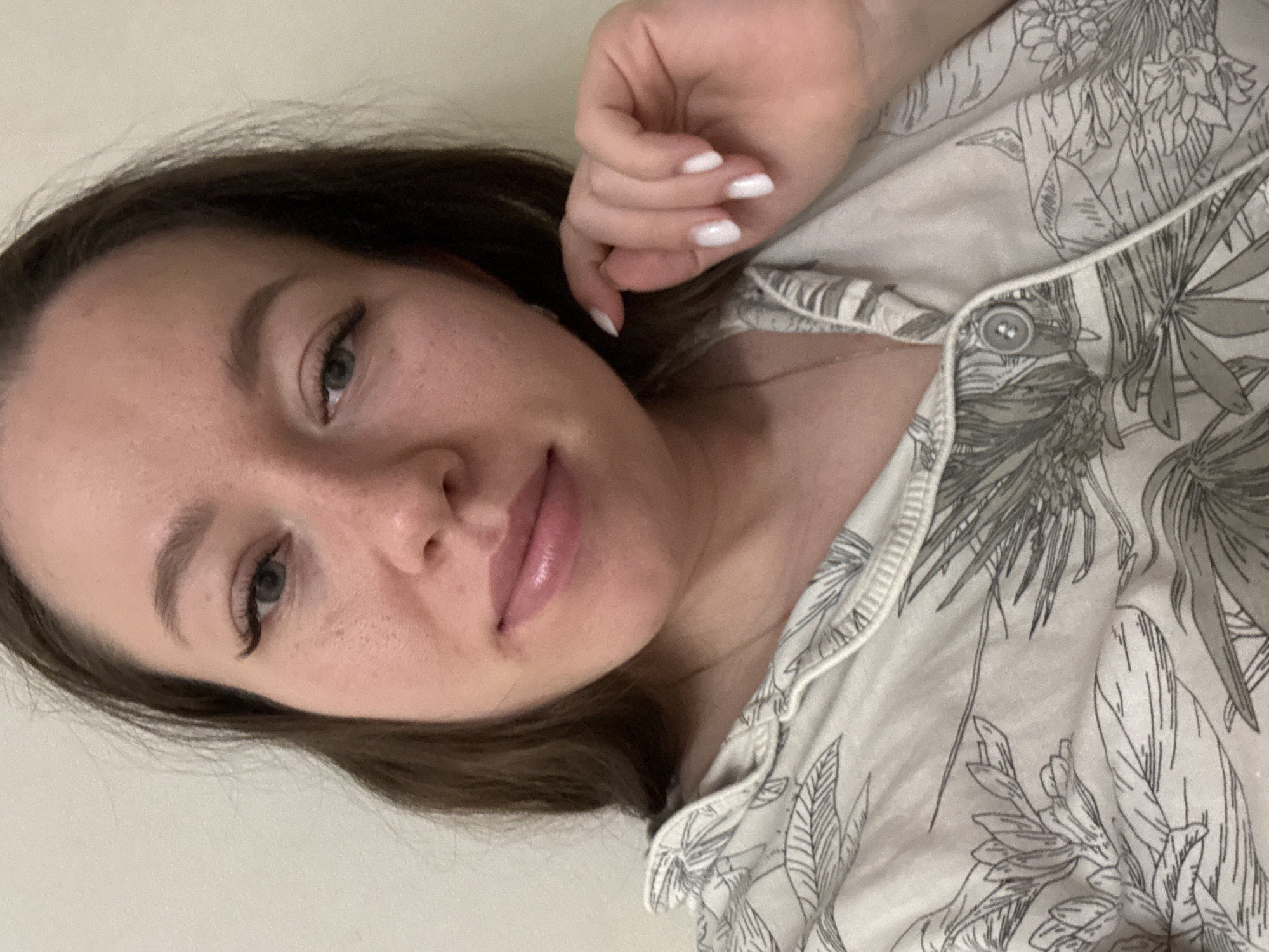 A woman with brown hair is lying down, likely indoors, wearing a light-colored button-down shirt with a floral pattern. Her nails are painted white. The location is presumed to be in the city of Ферма, Russia, but no details in the image confirm this.