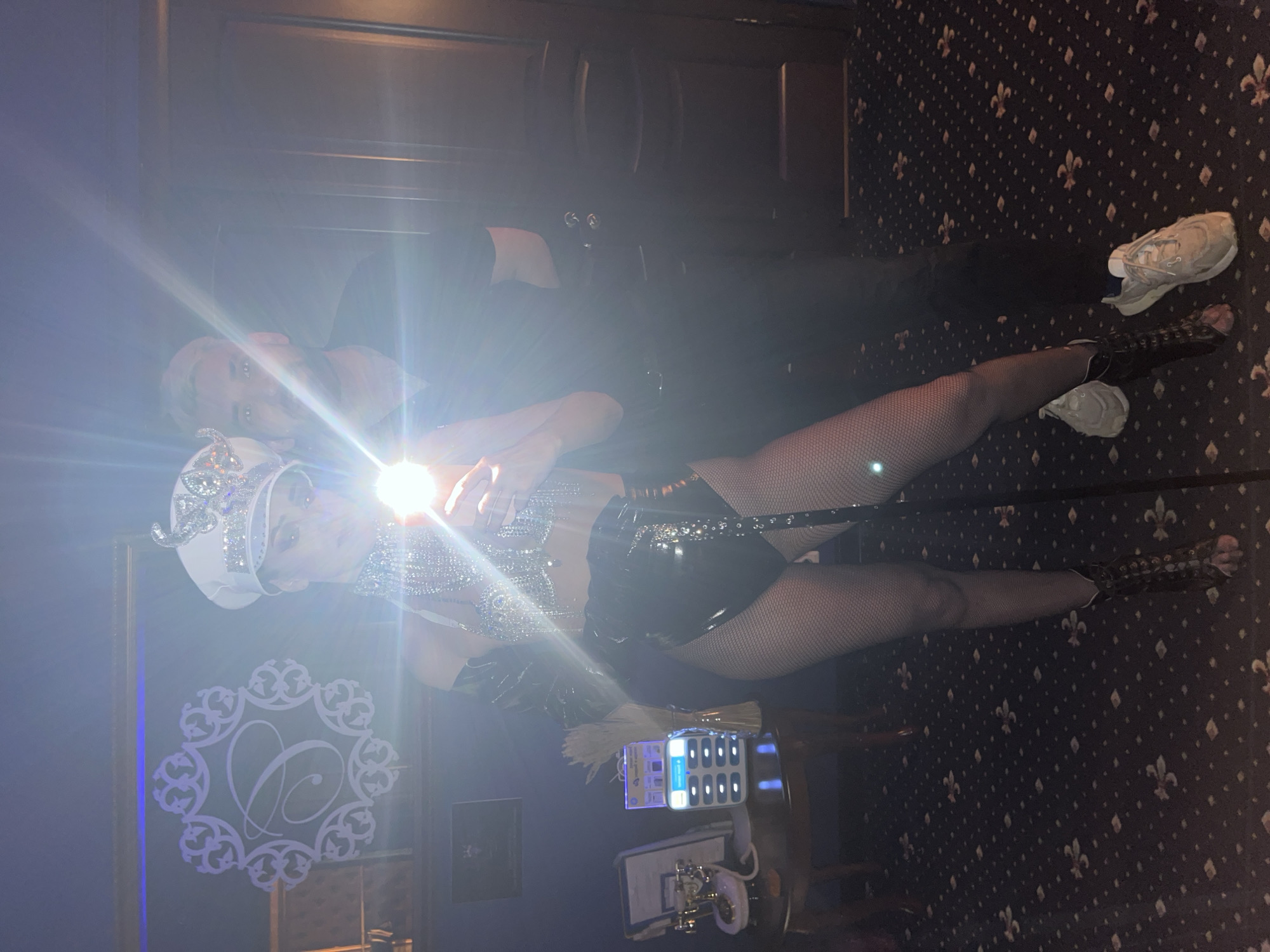 The image depicts two individuals in an dimly lit indoor environment, identified as Kazan, Russia. A strong camera flash originates from the center-left, creating significant lens flare.
On the left, a person, likely female, is dressed in elaborate performance attire. She wears a white military-style hat adorned with silver embellishments and a butterfly motif. Her upper body is covered by a sparkling, rhinestone-encrusted bra-top. She has a feather accessory on her left shoulder and wears shiny black, high-waisted shorts made of a material resembling latex or PVC. Her legs are covered in fishnet stockings, and she wears black, open-toed, high-heeled boots with strap details. She is holding a smartphone in her right hand, which is emitting the flash illuminating the scene.
To the right and slightly behind, a second person, likely male, stands. He has light hair and a beard, and wears a dark (appears black) short-sleeved collared shirt and dark trousers. His footwear consists of light-colored athletic sneakers. Both individuals are looking towards the camera.
The background on the left features a dark blue or purple wall with a framed mirror or artwork. Within this frame, a white illuminated emblem displays an ornate letter "B" or "P" surrounded by decorative swirls. Below this, a dark wooden table holds an antique-style rotary telephone (gold and white), a document stand, a white charging station with multiple illuminated blue slots, a white feather decoration, and a gold tassel. On the right, a dark wooden door with two silver door handles is visible. The floor is covered with a dark carpet featuring a repeating light-colored floral or heraldic pattern.