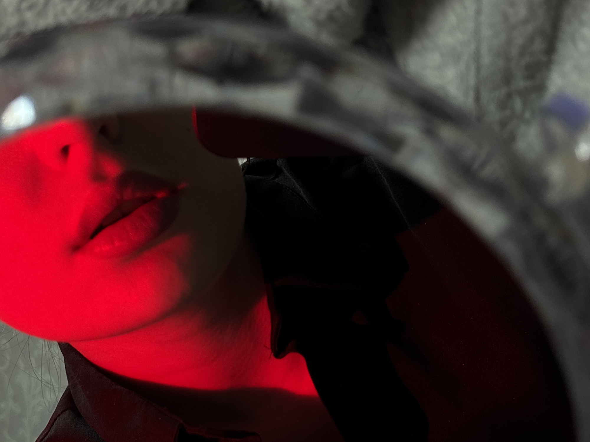 A close-up photograph features a person's face reflected in a distorted, circular surface, likely a mirror or lens. The left side of the face, including the lips, chin, and a portion of the cheek, is illuminated by a bright red light, creating a stark contrast against the darker areas. A black garment, possibly a shirt or jacket, is visible in the lower part of the reflection. The background, visible outside the circular reflecting surface, appears textured, potentially fabric. The image does not obviously reveal its location as Chaikovsky, Russia.