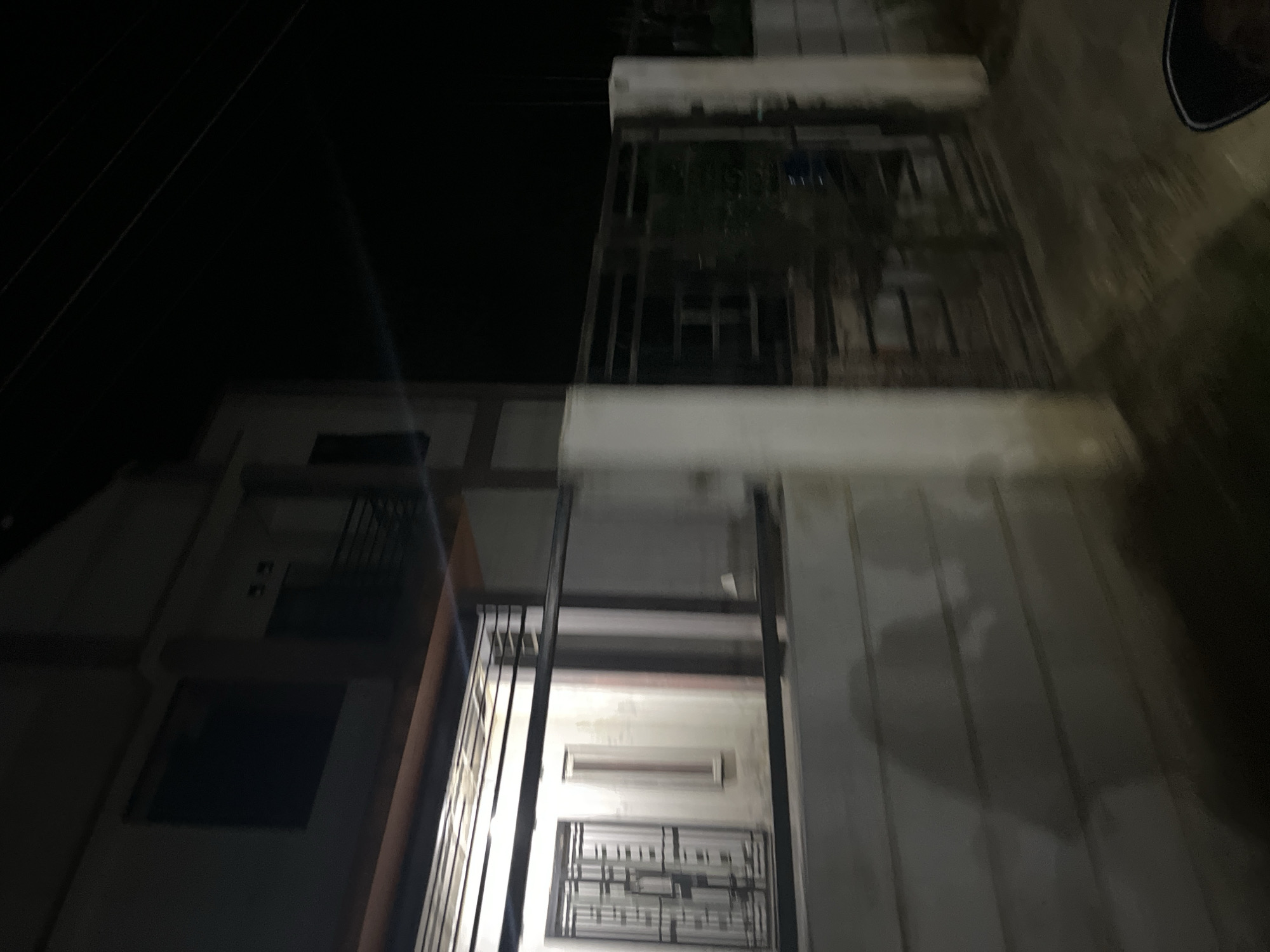 This media file depicts a nighttime scene in Menanggini, Indonesia, featuring a multi-story building with exterior metal railings and what appears to be a balcony or veranda. The building's facade is illuminated, contrasting with the dark night sky. Power lines are visible against the dark background. The structure suggests residential or commercial use. There are no people or specific activities clearly discernible, but the overall impression is one of a quiet, possibly deserted, urban or semi-urban environment at night. The low lighting and angles obscure many finer details, but the architectural elements of the building, such as windows and railings, are the primary focus. The time of day is clearly night, and the weather appears to be clear, as evidenced by the unobstructed view of the sky. No text is visible in the image.
