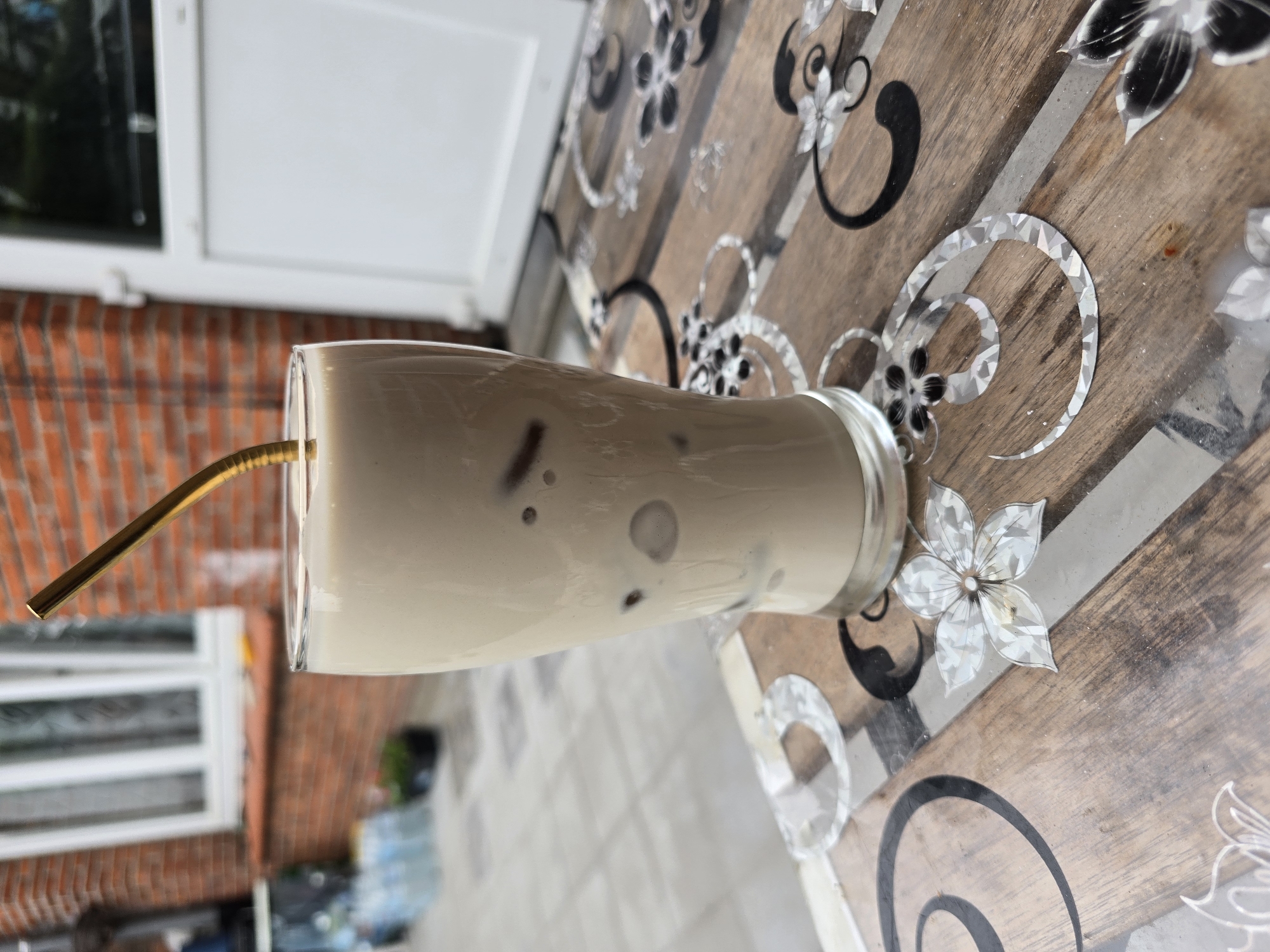 A tall glass of iced coffee with a golden metal straw is placed on a wooden table with a decorative pattern. The table is outdoors, with a brick building and a white door in the background. The coffee is a light brown color with ice cubes visible inside. The straw is angled out of the glass. The scene is in Ghent, Belgium, specifically in the area of the Vrijdagsmarkt square, known for its outdoor cafes and brick architecture.