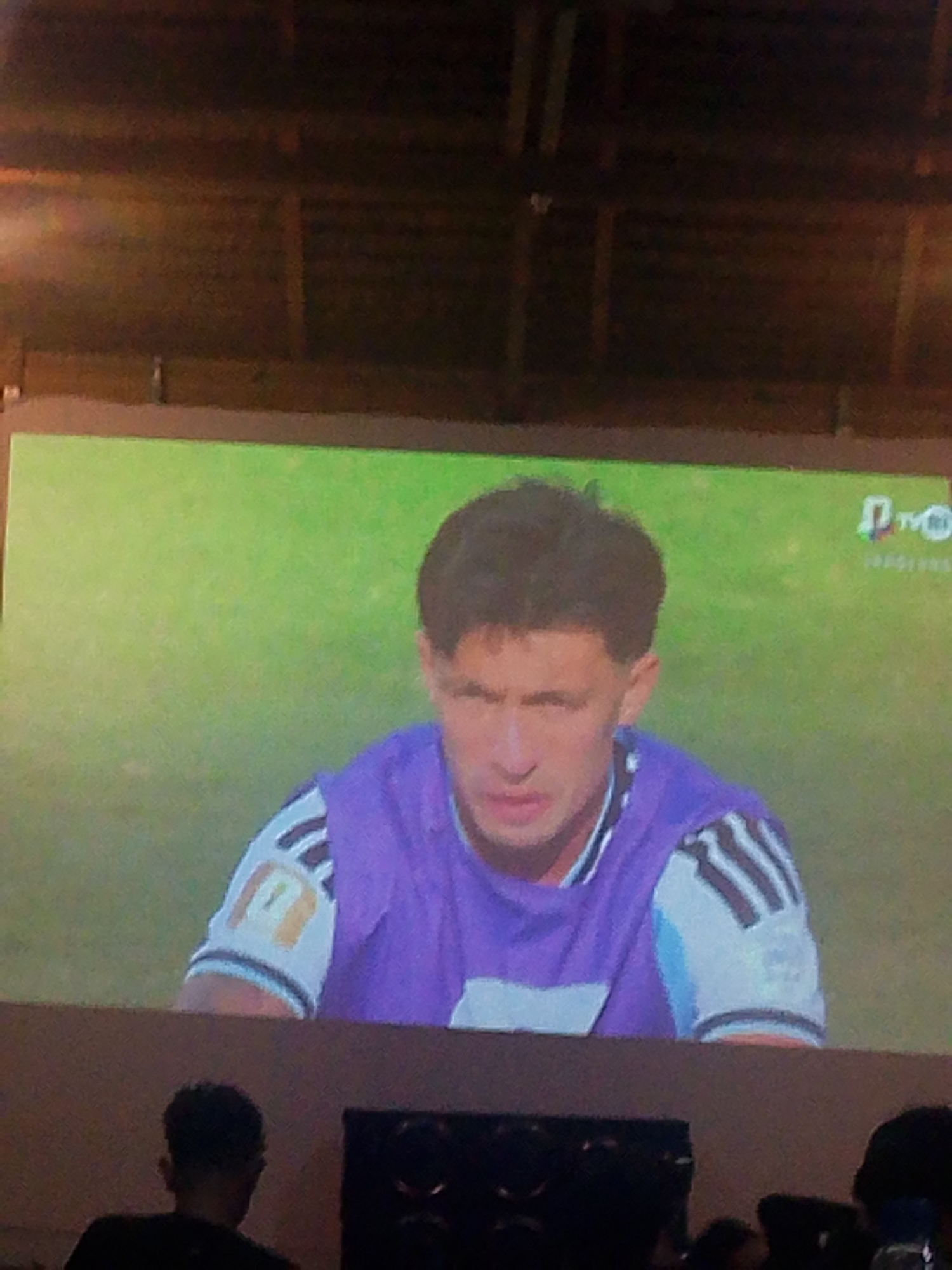 A video plays on a large screen, displaying a close-up of a soccer player wearing a purple and white jersey. The player has dark hair and is looking down, with a neutral expression. The background of the video is a blurred green field. Below the screen, silhouettes of people are visible. The scene appears to be in a public indoor space, possibly a restaurant or bar, given the wooden ceiling structure and the presence of what looks like other screens or televisions in the foreground. The location is identified as Meunasahmee, Indonesia.