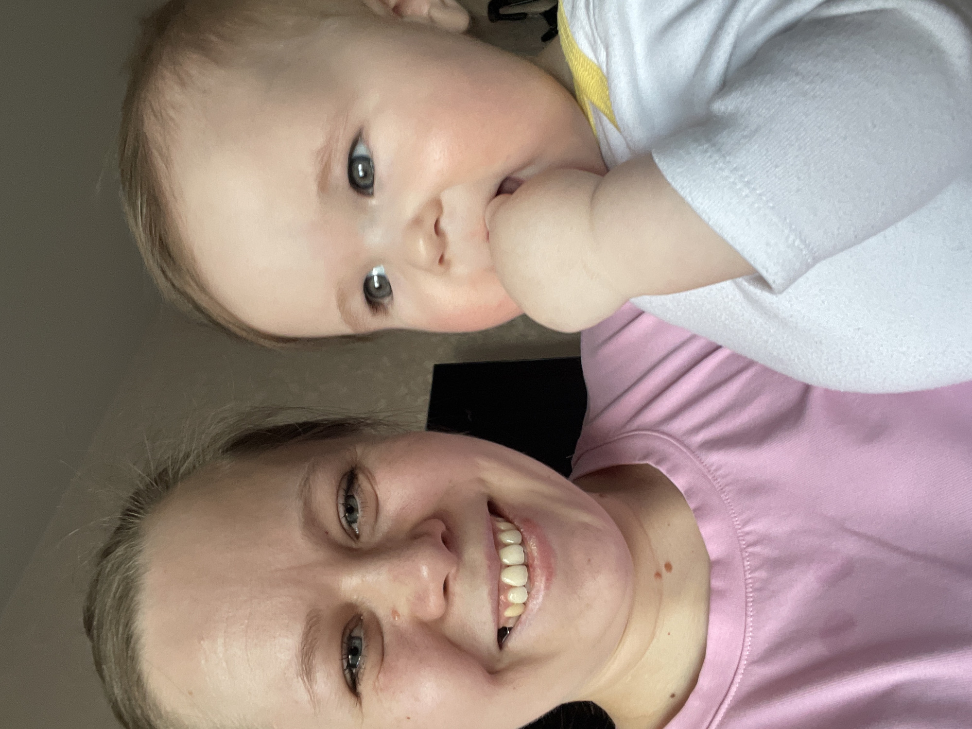 A selfie-style image shows a woman and a baby. The woman has fair skin, blonde hair, and is wearing a pink shirt. She is smiling at the camera. The baby is above her, wearing a white shirt with a yellow collar, and sucking on their fingers. The background is blurry and indistinct, suggesting an indoor setting. The image does not provide any information about the location in the city of Chaikovsky, Russia.