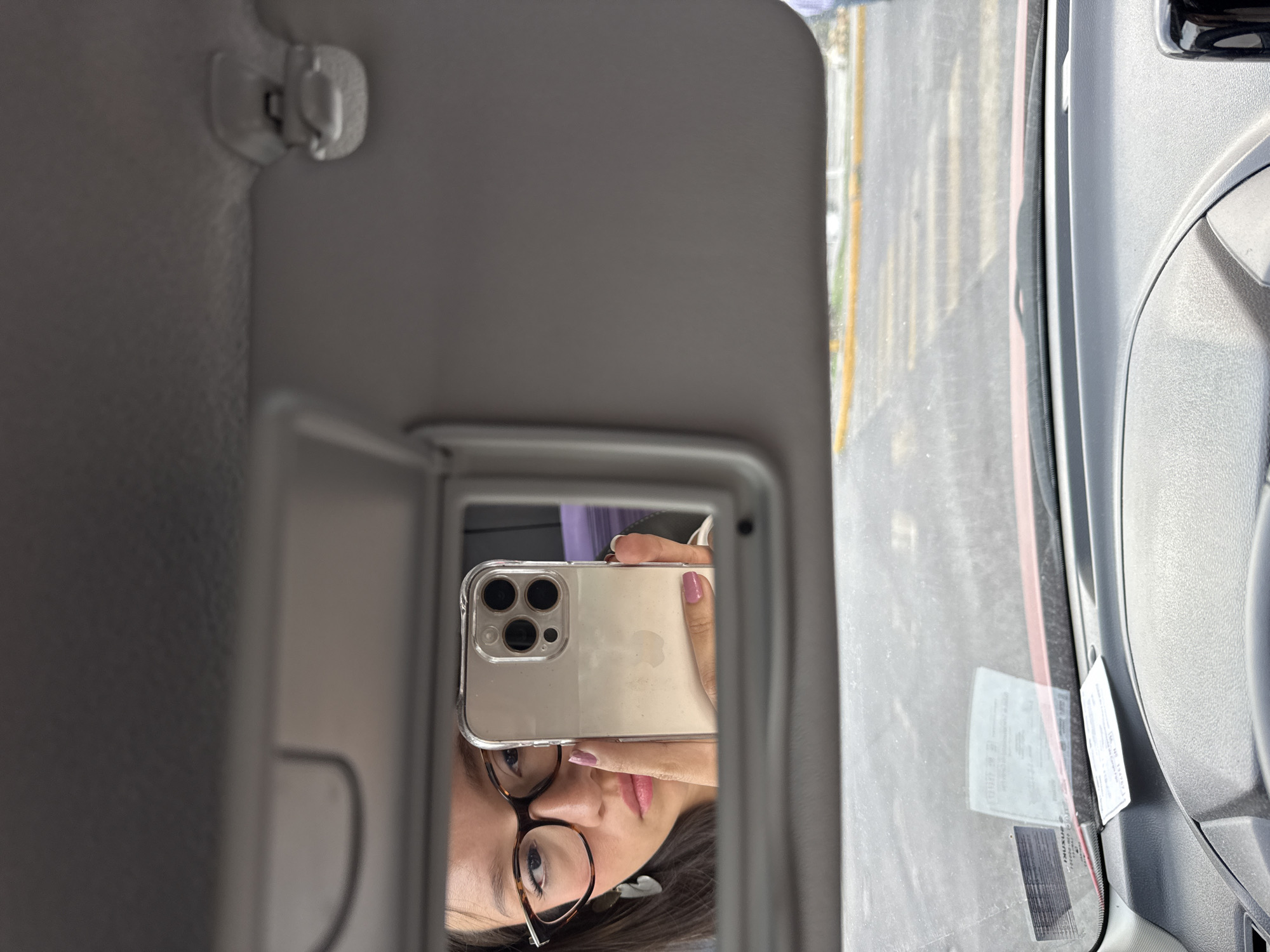 A person takes a selfie in a car mirror. The phone has a clear case. The person wears glasses and has pink painted nails. A road is visible through the windshield. The location cannot be determined from the image.