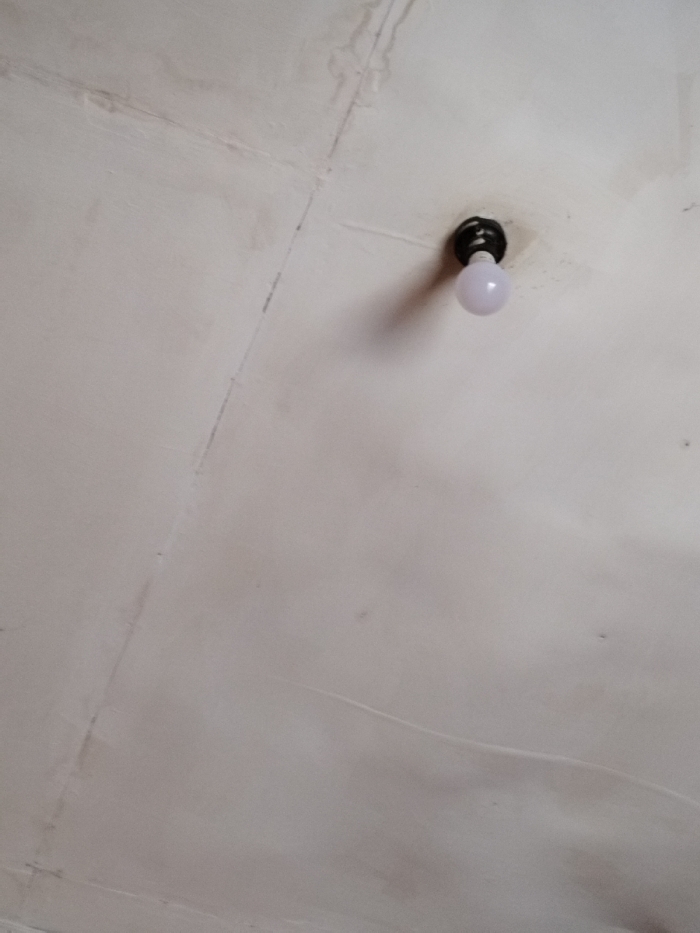 An upward-angle photograph depicts an interior ceiling surface in Muka, Indonesia. A single, spherical, opaque white light bulb is affixed to a dark, circular ceiling-mounted fixture, positioned in the upper-right quadrant of the frame. The ceiling is a light, possibly off-white, color and displays extensive discoloration, dark streaks, and irregular blotches consistent with moisture damage or mold. Visible seams and hairline cracks are present across the ceiling surface, particularly prominent in the upper-left and central portions. No individuals, actions, or interactions are visible. The light bulb appears unlit.