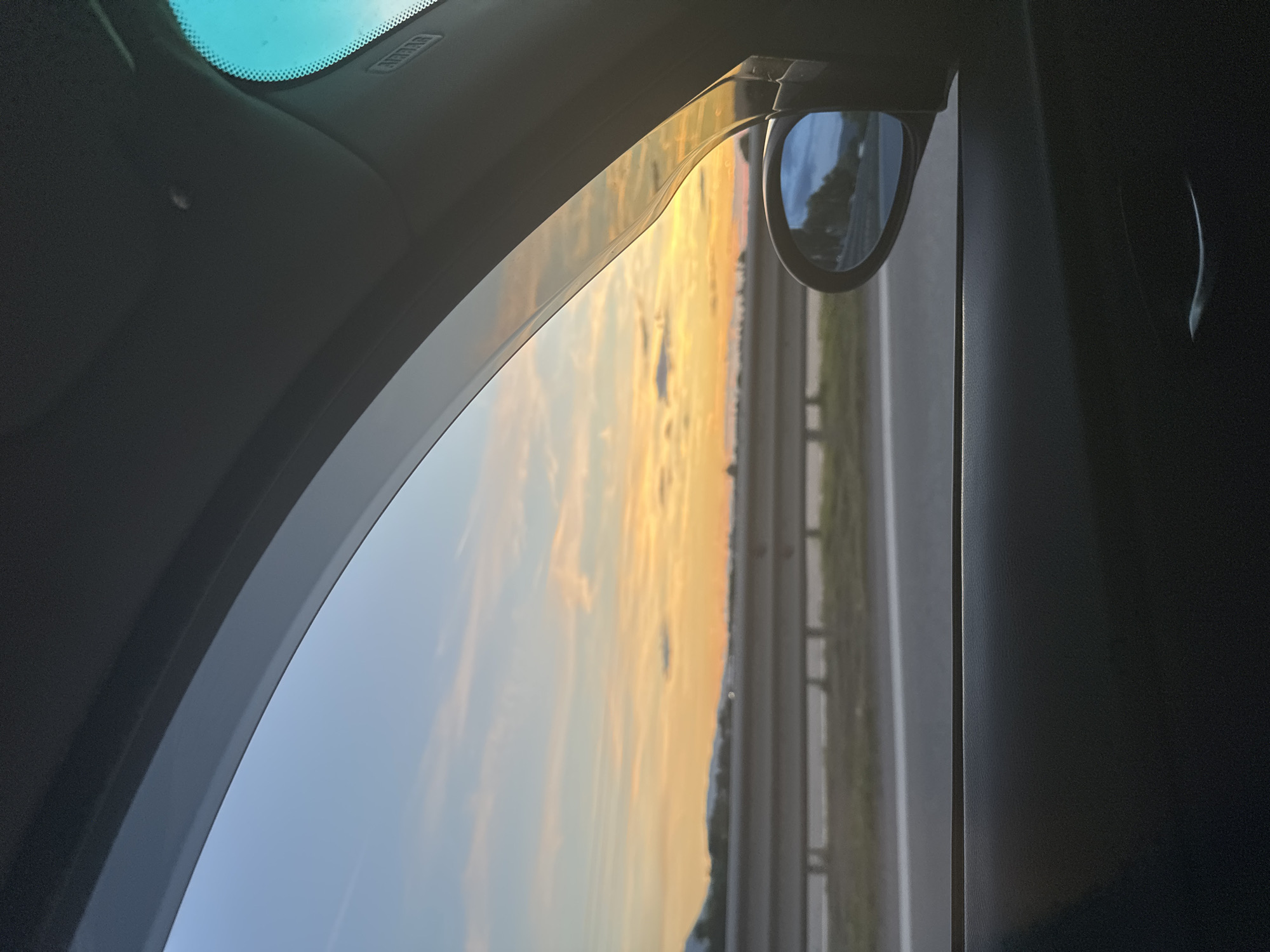 The image, taken from inside a car, shows a sunset view through the passenger window. The sky is a gradient of blue, orange, and yellow, with scattered clouds. A highway barrier and trees are visible in the distance. A side mirror reflects a portion of the scenery. The interior of the car, including the door panel and window trim, is dark and out of focus. The location is identified as Krivina, Bulgaria.