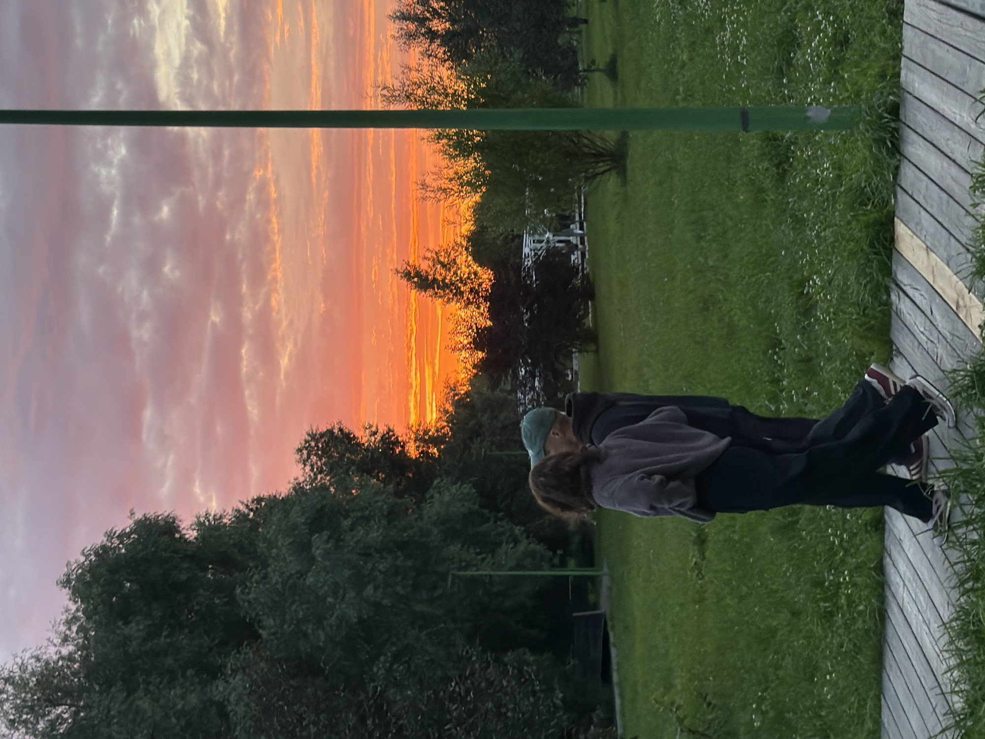 Two people are standing on a wooden walkway, their backs to the camera, as they face a vibrant sunset. The sky is ablaze with hues of orange, pink, and yellow, with streaks of white clouds scattered across it. The setting appears to be a park or outdoor recreational area, with lush green grass and trees visible in the background. A green pole runs horizontally across the frame, partially obscuring the sky. The wooden walkway is made of light-colored planks and has a curved design. The time of day is clearly sunset, indicated by the dramatic colors in the sky. There are no visible signs of text in the image. The overall mood is peaceful and contemplative, with the subjects seemingly enjoying the natural beauty of the sunset. The location context of Kazan, Russia, suggests this scene is taking place in an urban park or a suburban outdoor space.