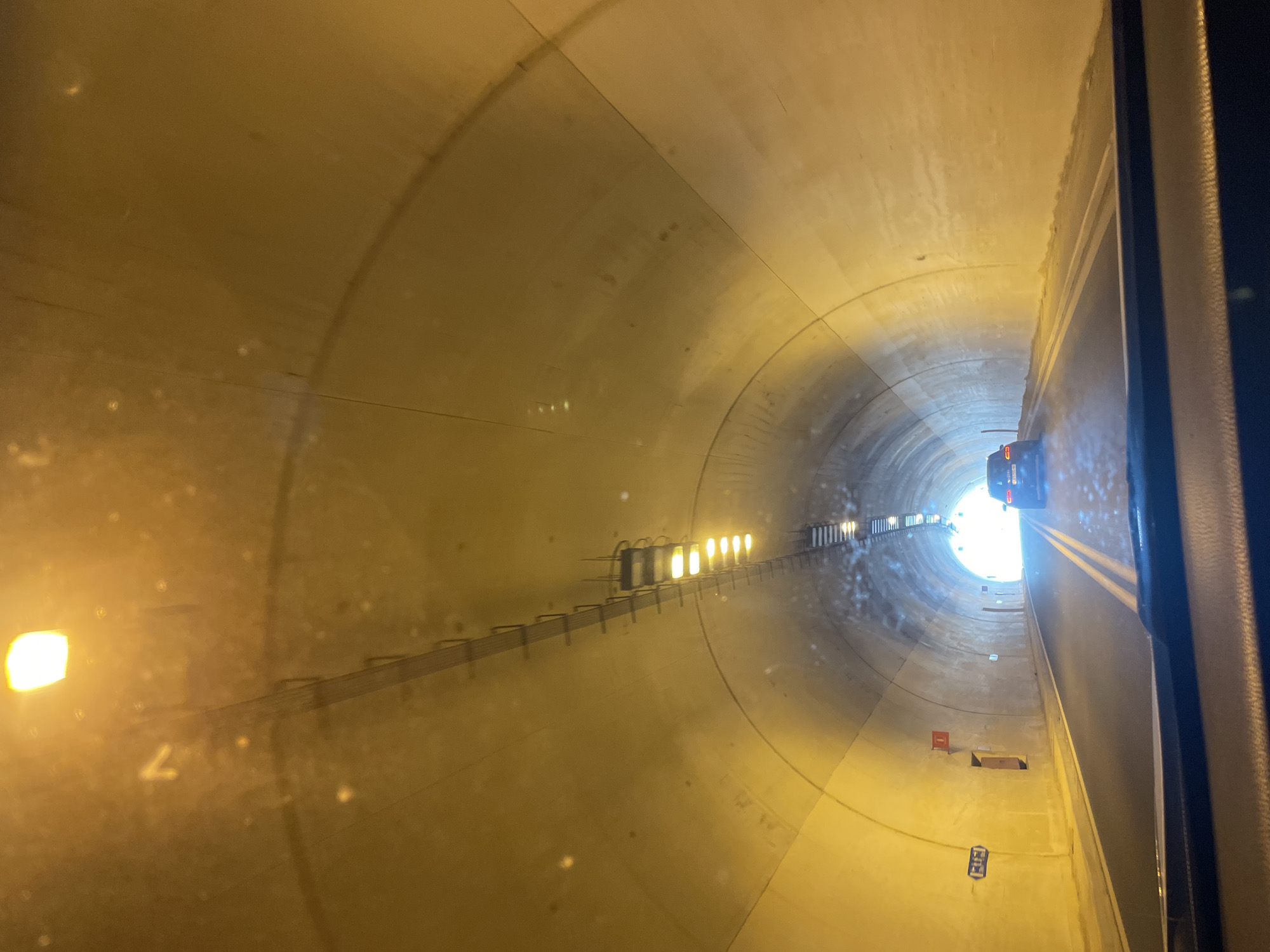 The image shows the interior of a vehicle looking out towards the end of a tunnel. The tunnel walls are illuminated by overhead lights, creating a yellow glow. A single car is visible ahead, driving towards the bright exit of the tunnel. There are lane markings on the road and some signage on the left wall of the tunnel. The overall scene depicts travel through an underground passage.