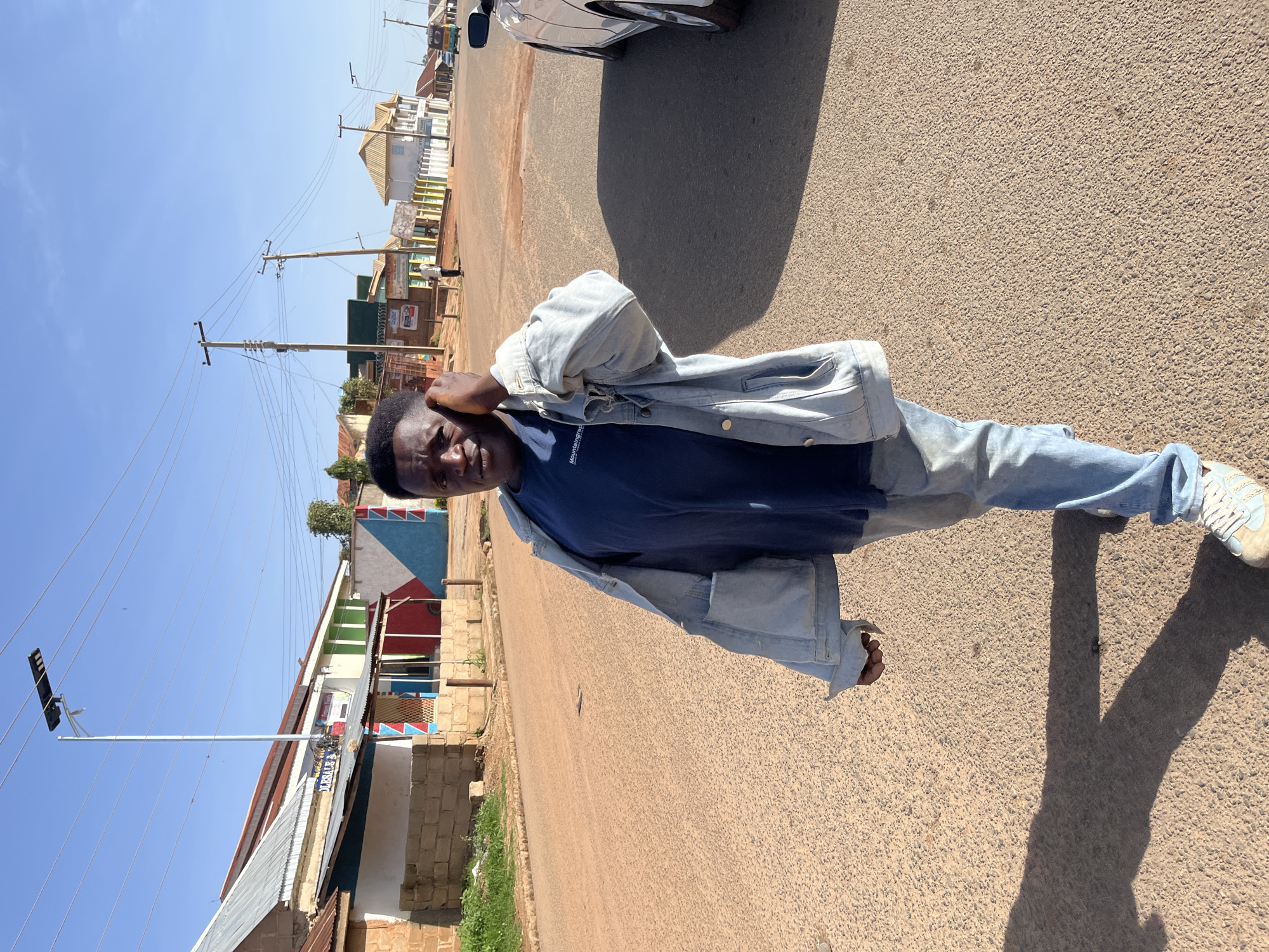 The image, originally rotated, depicts a young man standing on a paved road in what appears to be a town in Jos, Nigeria. The man, likely of Black ethnicity, is centrally positioned, looking directly at the camera with a neutral to slight smile. He is dressed in a dark blue t-shirt with white text partially visible as "Mountain" or "Mountaingrad" across the chest, a light blue denim jacket worn over it, light blue jeans, and light-colored sneakers with white laces. His left hand is raised towards his ear/head, while his right arm is by his side.
The road he stands on is light brown/grey, unlined, and stretches into the distance. On either side, various single and two-story buildings line the street, some featuring corrugated metal roofs and others tiled. Several buildings are painted in bright hues like red, blue, green, and white, showcasing local architecture. Numerous utility poles, both wooden and concrete, with a dense network of overhead wires, are prominent along the road; one pole features a solar-powered street light. A section of a silver or white car (front wheel and fender) is visible parked on the right side of the road.
Long shadows, including that of the man, are cast on the road, suggesting either early morning or late afternoon sunlight. The sky is clear and bright blue, indicating sunny weather. The overall scene conveys the atmosphere of an active urban or semi-urban environment.