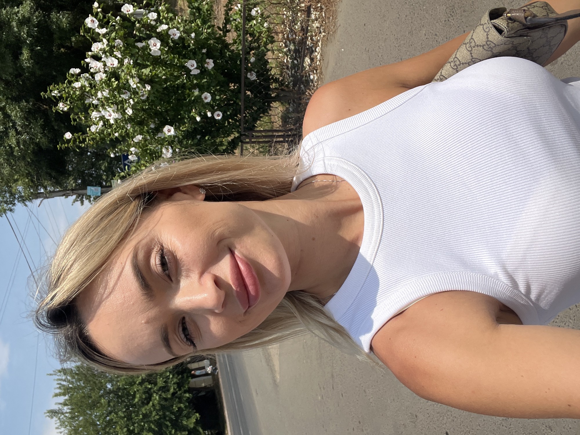 A blonde woman in a white ribbed tank top smiles at the camera in a selfie. She is outdoors, with trees and a bush with white flowers in the background. A road is visible to her left, and a fence is behind the bush. She is carrying a light brown handbag with a Gucci logo. The image appears to be taken during the daytime, as indicated by the bright sunlight and clear sky. The location is identified as Fontanka, Ukraine.